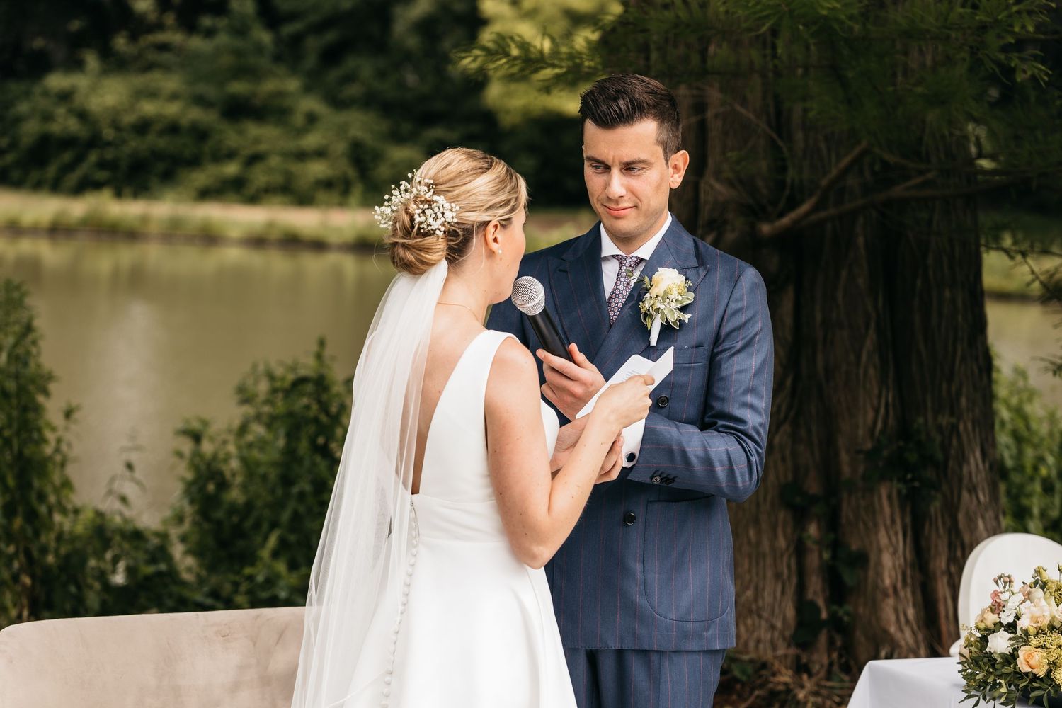 Groom in navy suit reads vows to bride in white dress during outdoor lakeside wedding ceremony.