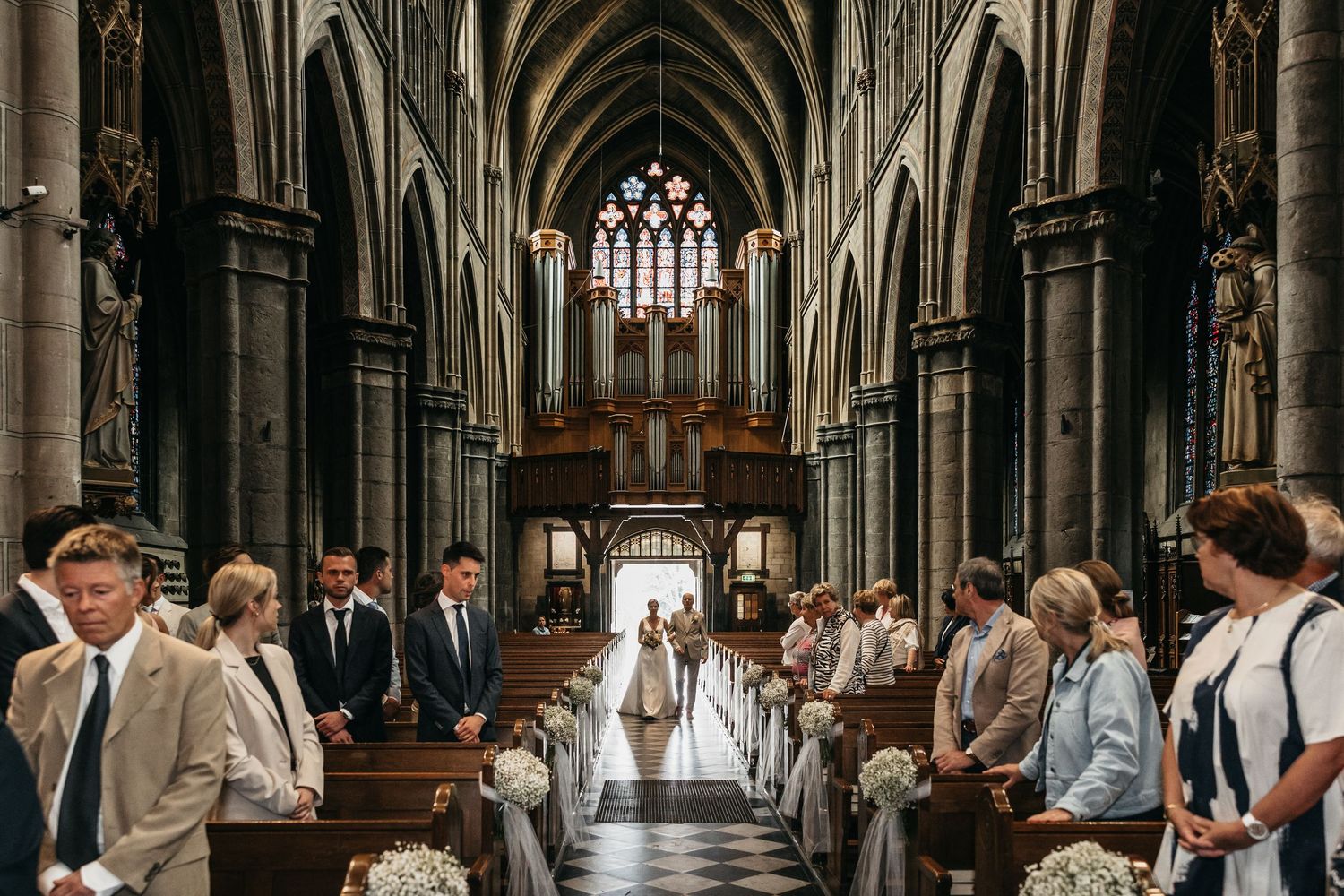Wedding ceremony inside grand Gothic church with guests lining the aisle as bride walks in.