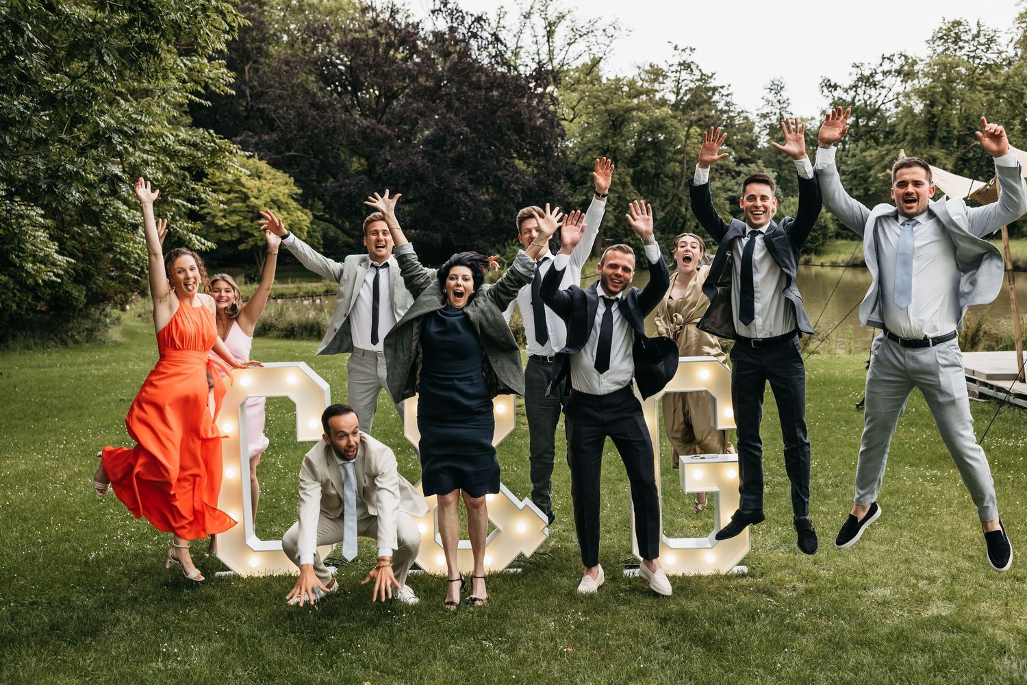 Wedding party jumping joyfully outdoors near large decorative letters on green lawn.