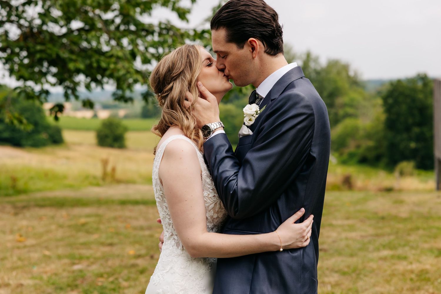 Bride and groom sharing a romantic kiss outdoors with green fields and trees in the background.