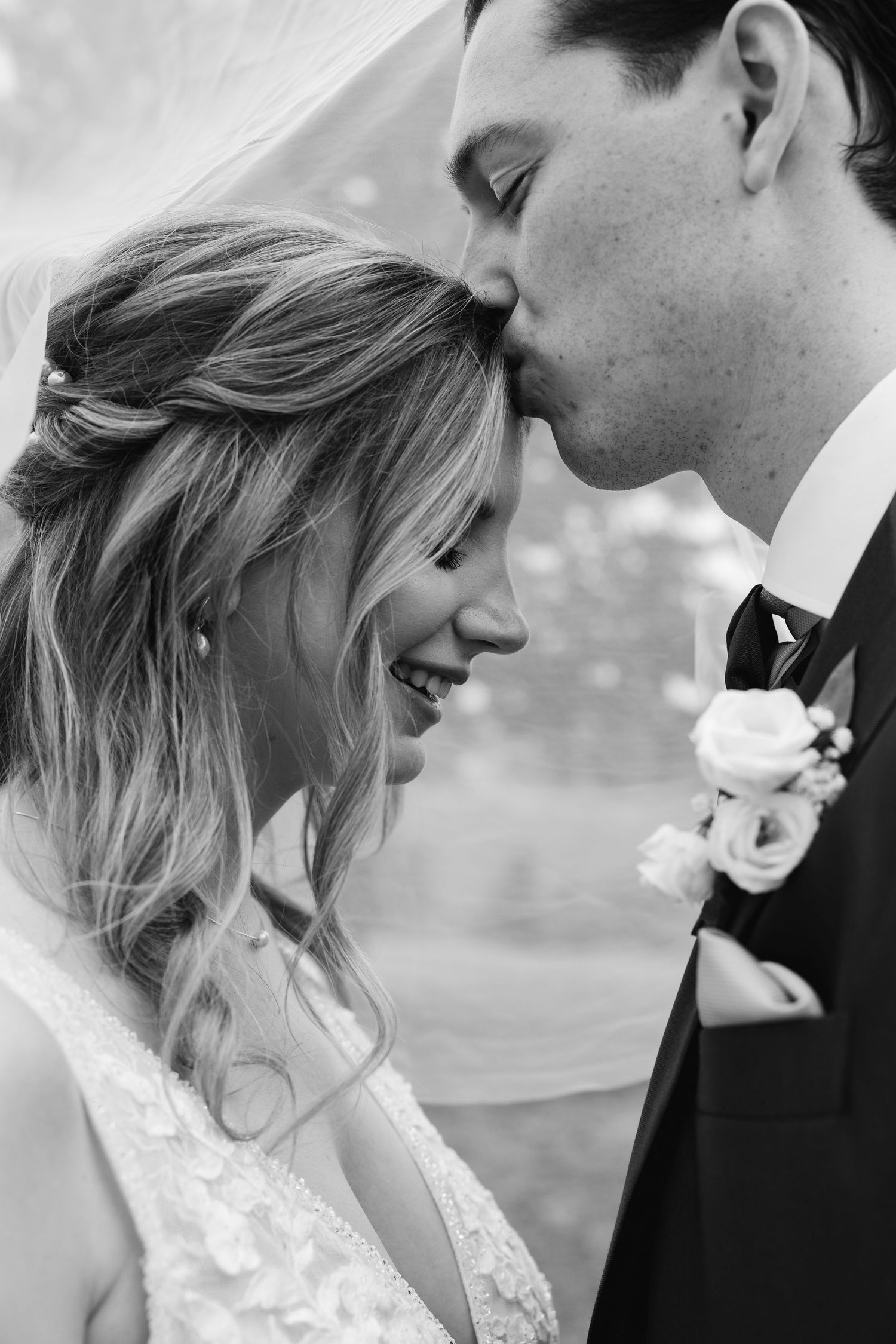 Black and white portrait of groom kissing brides forehead as she laughs joyfully beneath a veil.