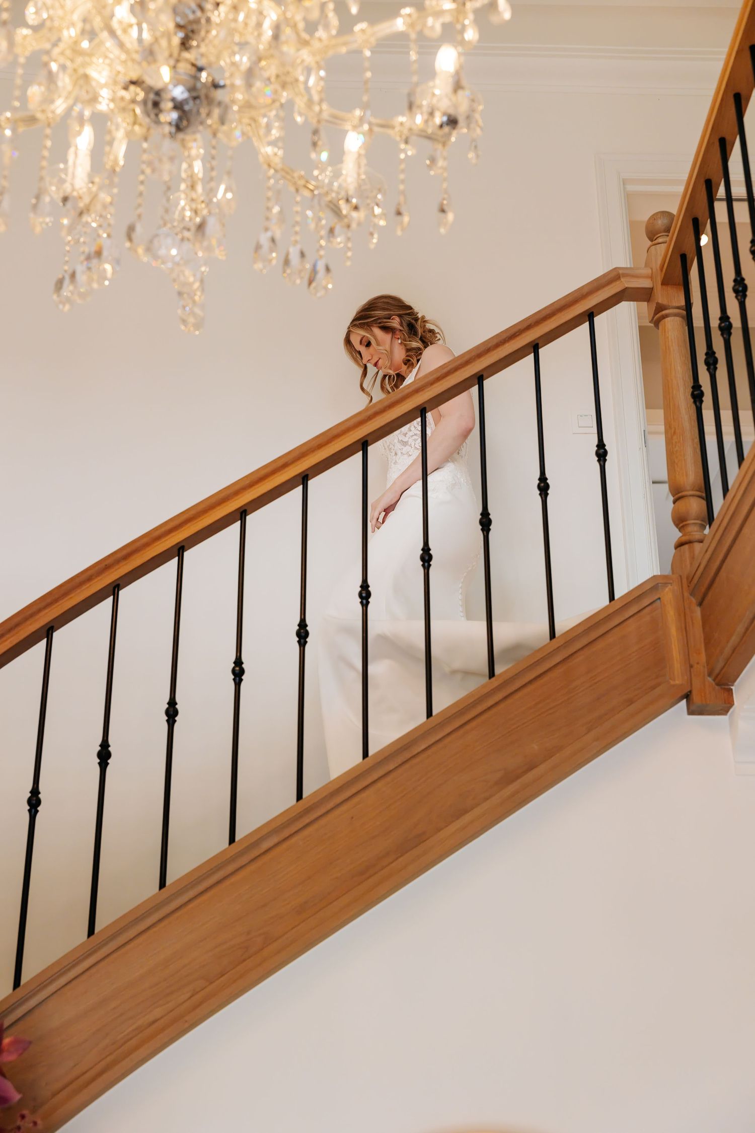 Bride in white gown peeks over a wooden banister while descending a staircase under a crystal chandelier.