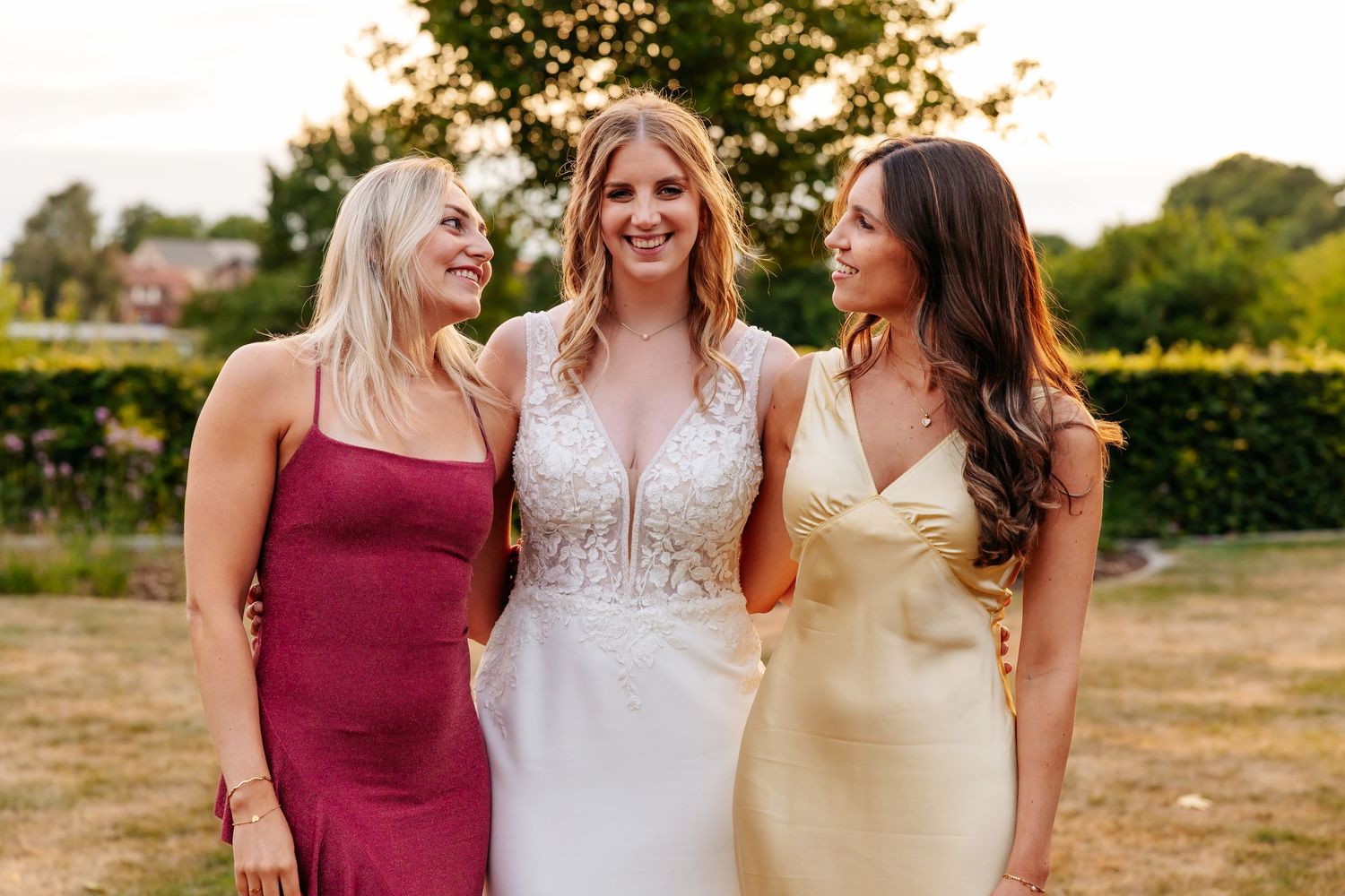 Bride poses with two bridesmaids in burgundy and gold dresses at an outdoor wedding.