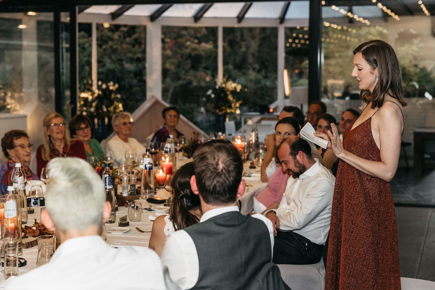 Woman in rust dress delivering a heartfelt wedding speech to seated reception guests.