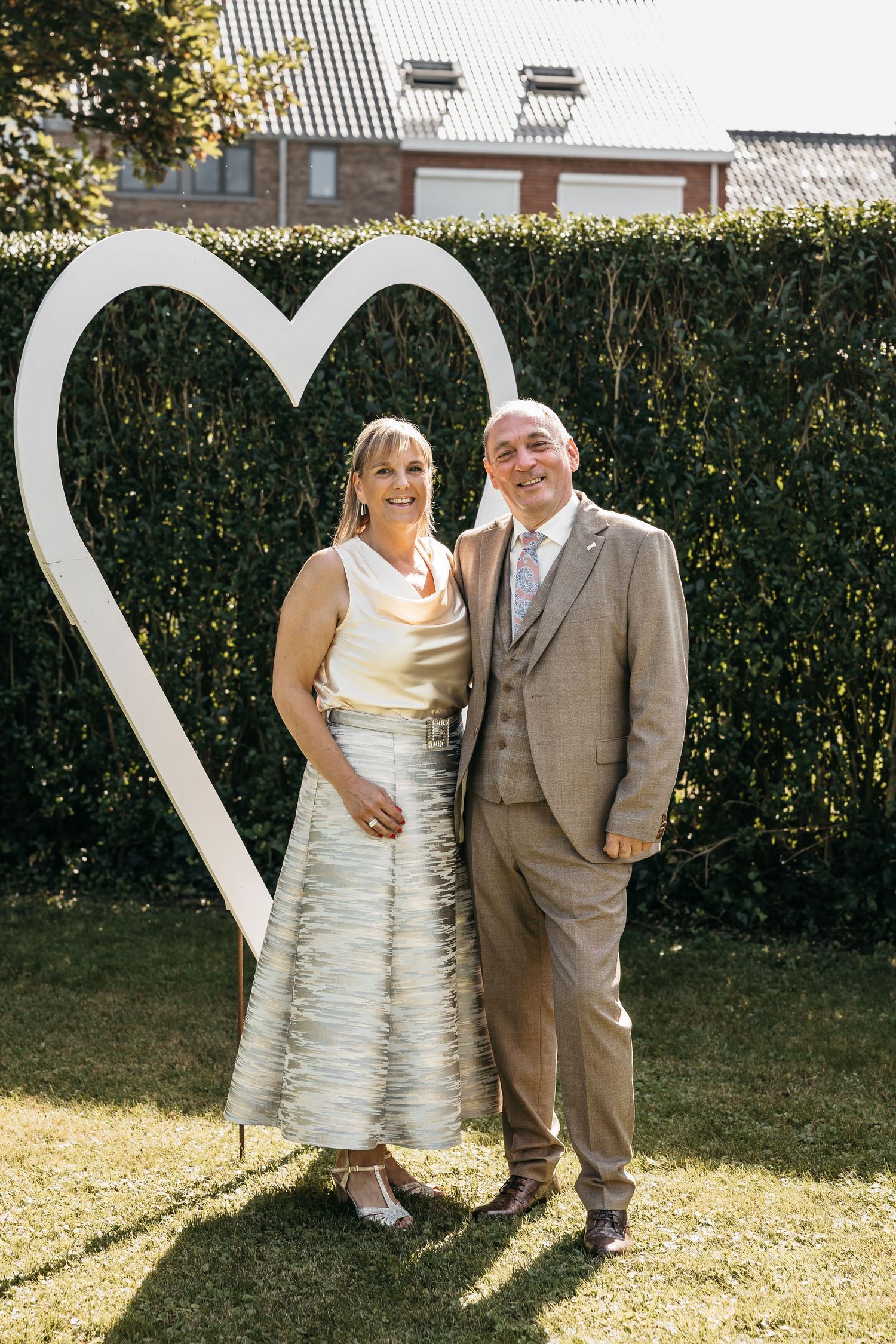 Smiling couple poses in front of a large white heart sculpture in a sunny garden at a wedding.