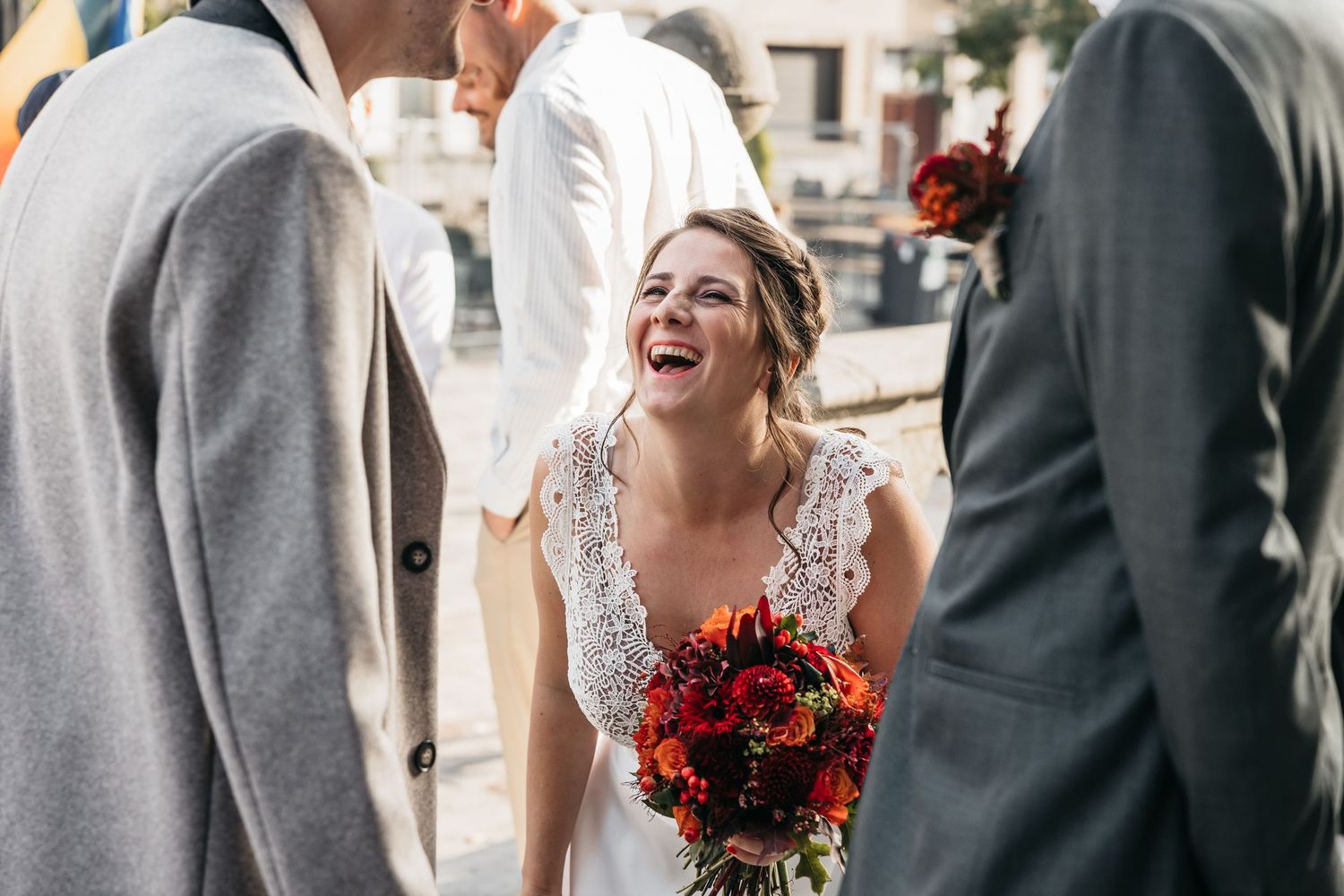 Laughing bride in beaded white gown holding red autumn bouquet surrounded by wedding guests outdoors.