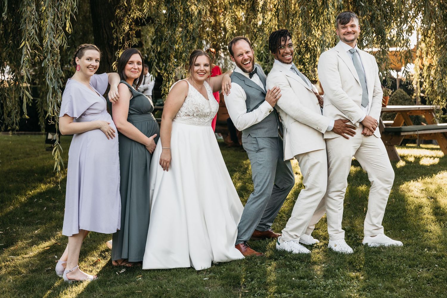 Bride and groom posing joyfully with wedding party guests under a willow tree outdoors.