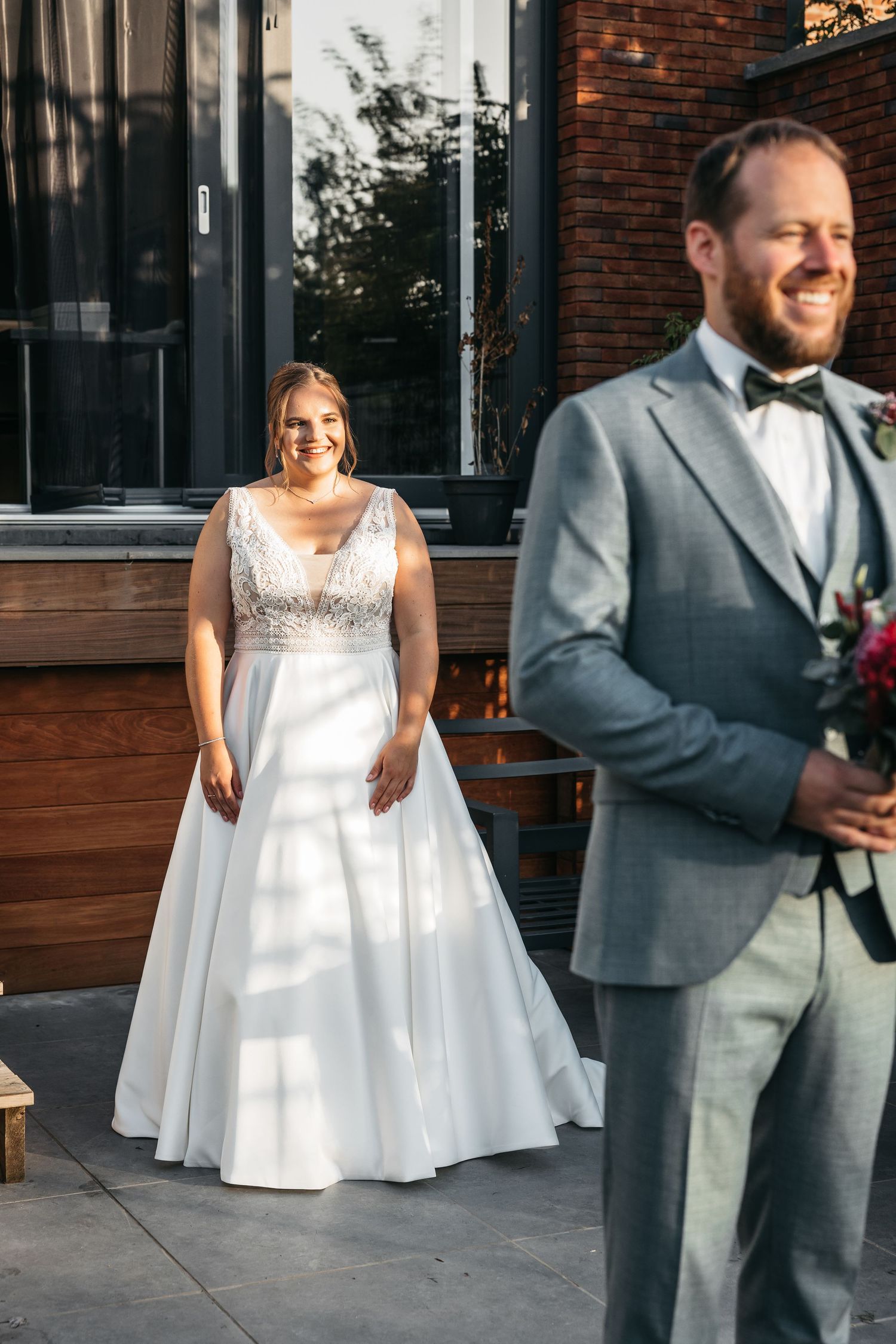 Smiling bride in white lace gown stands behind laughing groom holding a bridal bouquet.
