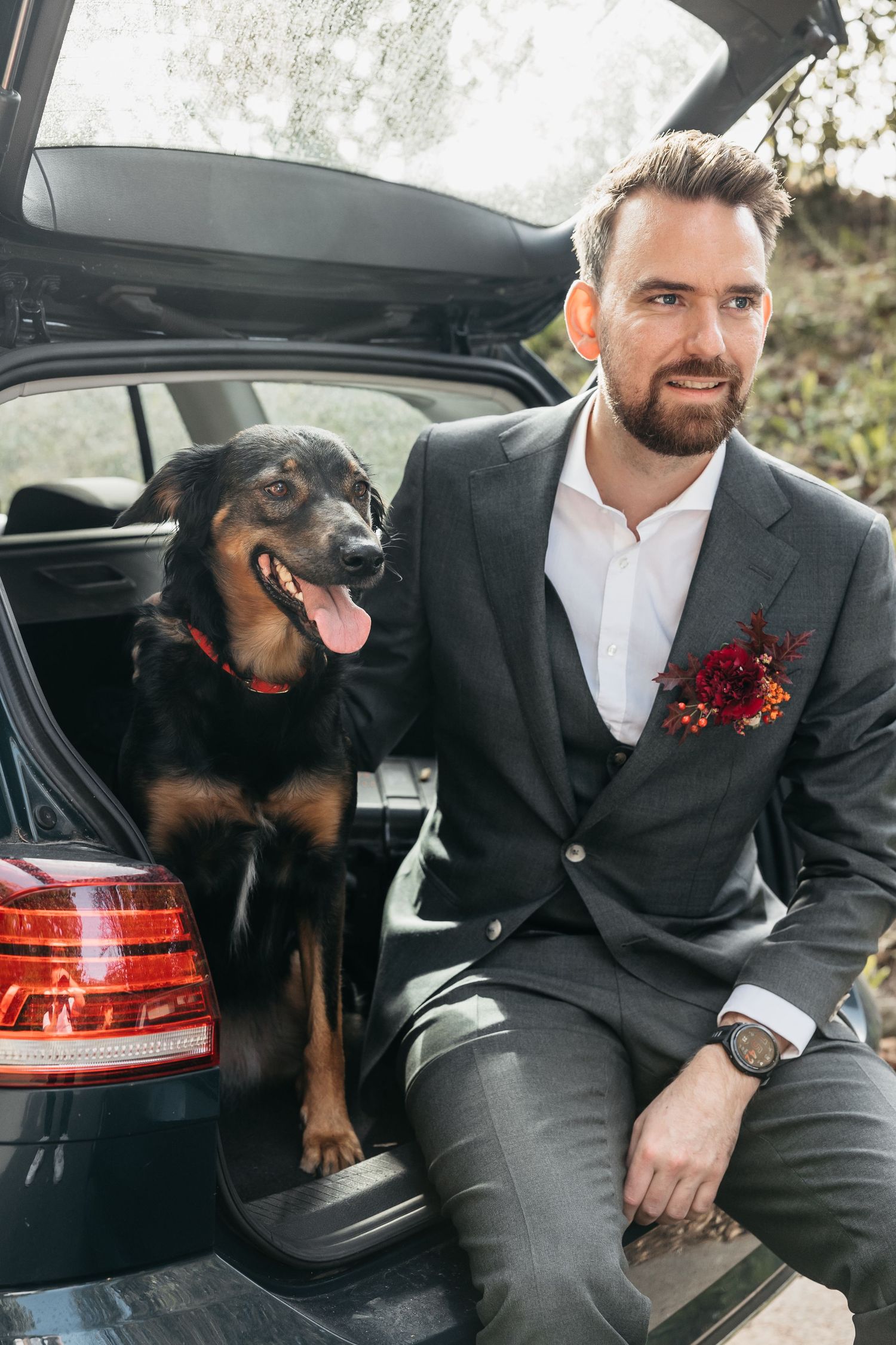 Groom in grey suit with floral boutonniere sitting in car trunk with black dog.
