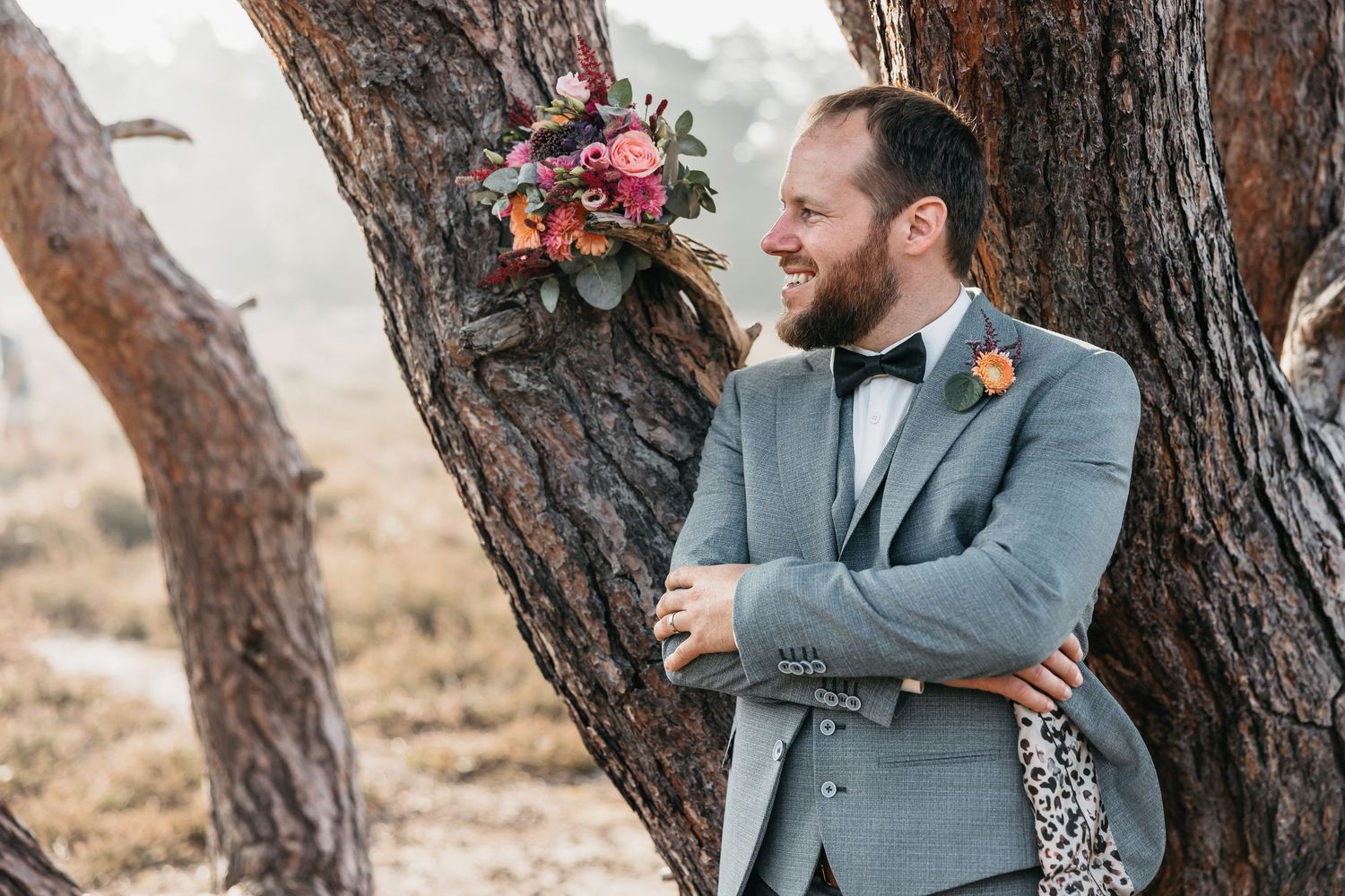 Groom in grey wedding suit with bow tie leans against a pine tree smiling beside a vibrant bridal bouquet.