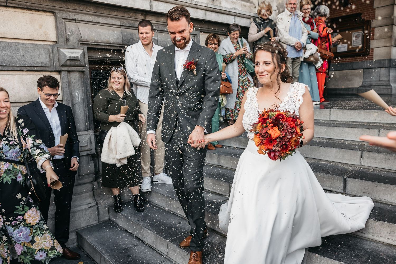 Newlyweds laugh joyfully as guests shower them with confetti on steps of elegant wedding venue.