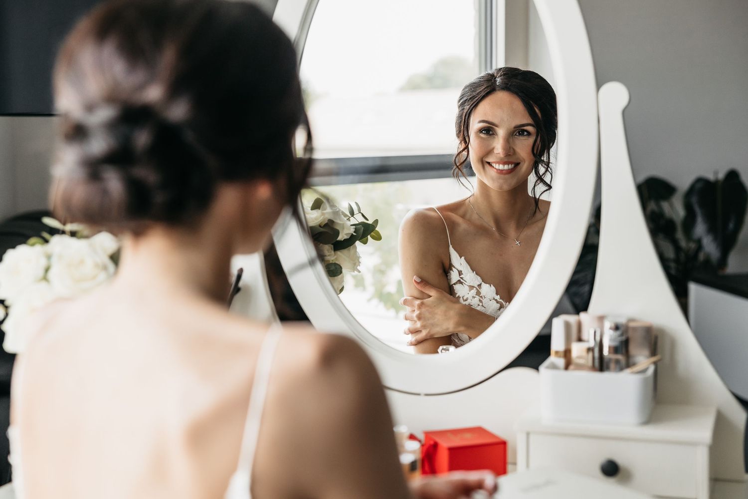 Radiant bride in white lace gown smiles at her reflection in a vintage oval mirror at a vanity table.