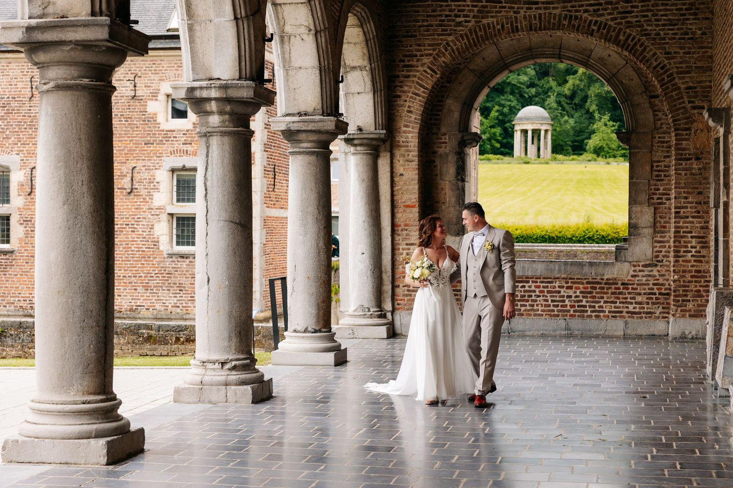 Bride and groom share a romantic moment under stone arches of a historic brick building with lush green gardens beyond.