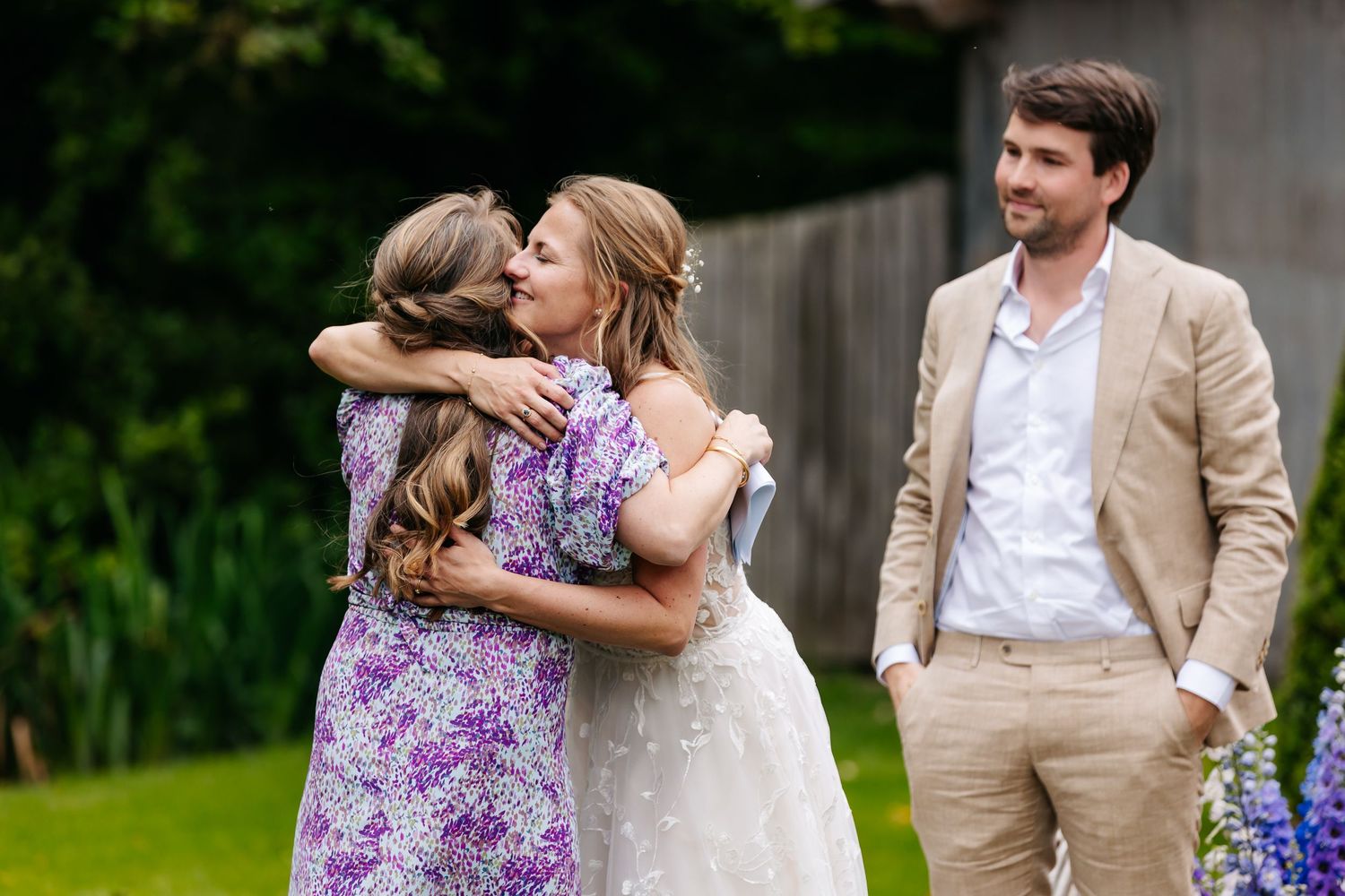 Bride in white gown hugs guest in purple floral dress while groom watches at outdoor wedding.