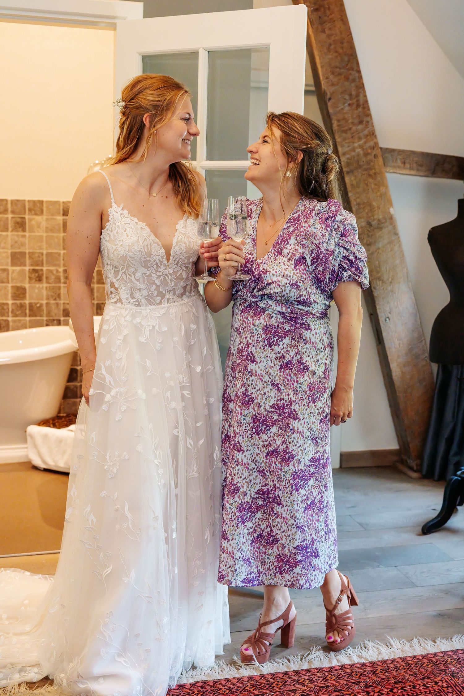 Bride in white lace gown toasting champagne with woman in floral dress during wedding preparations indoors.
