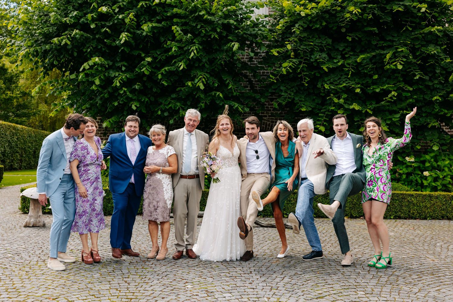 Joyful wedding party group photo with bride and groom surrounded by family and friends outdoors.