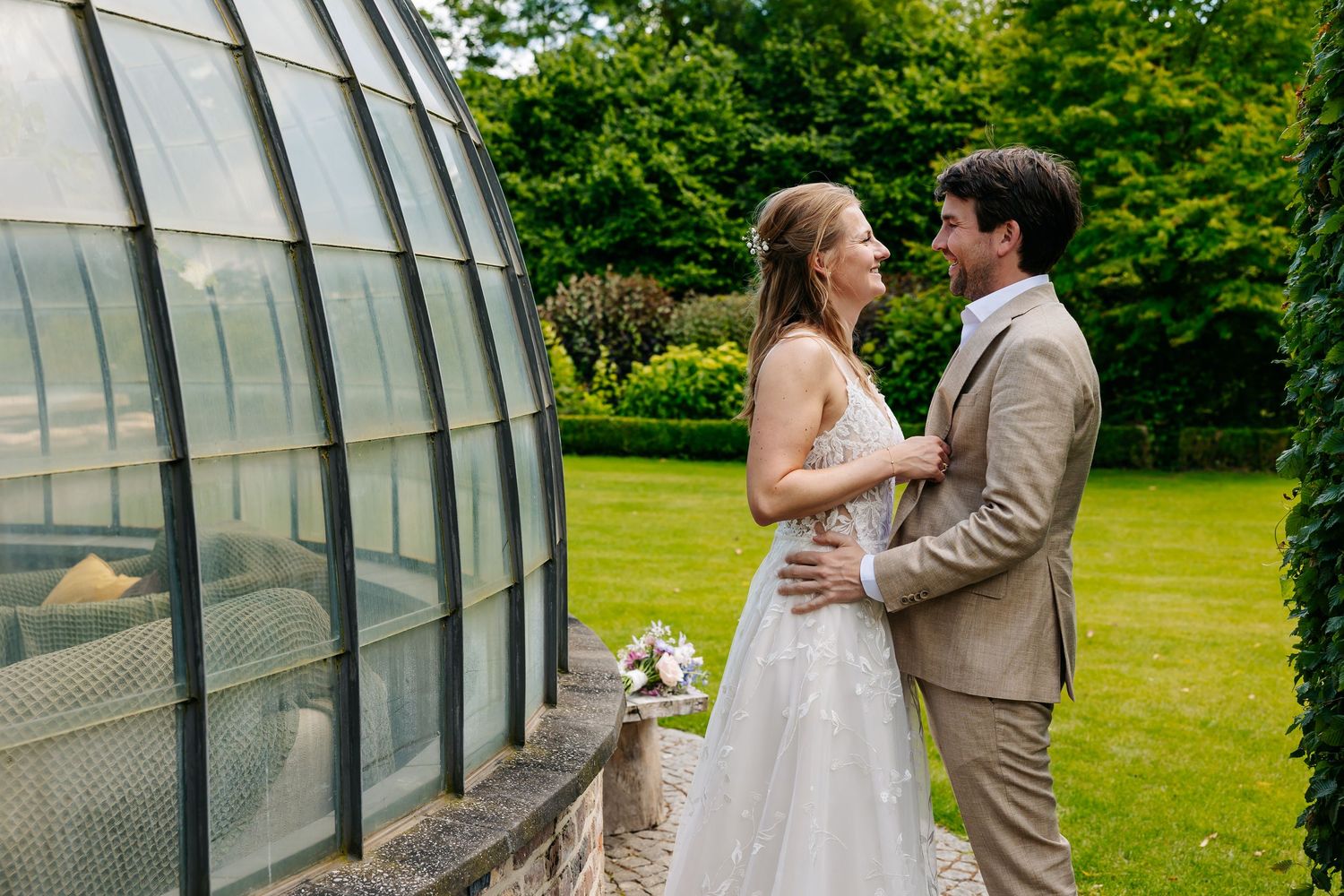 Bride and groom sharing a romantic moment beside a glass greenhouse in a lush green garden.