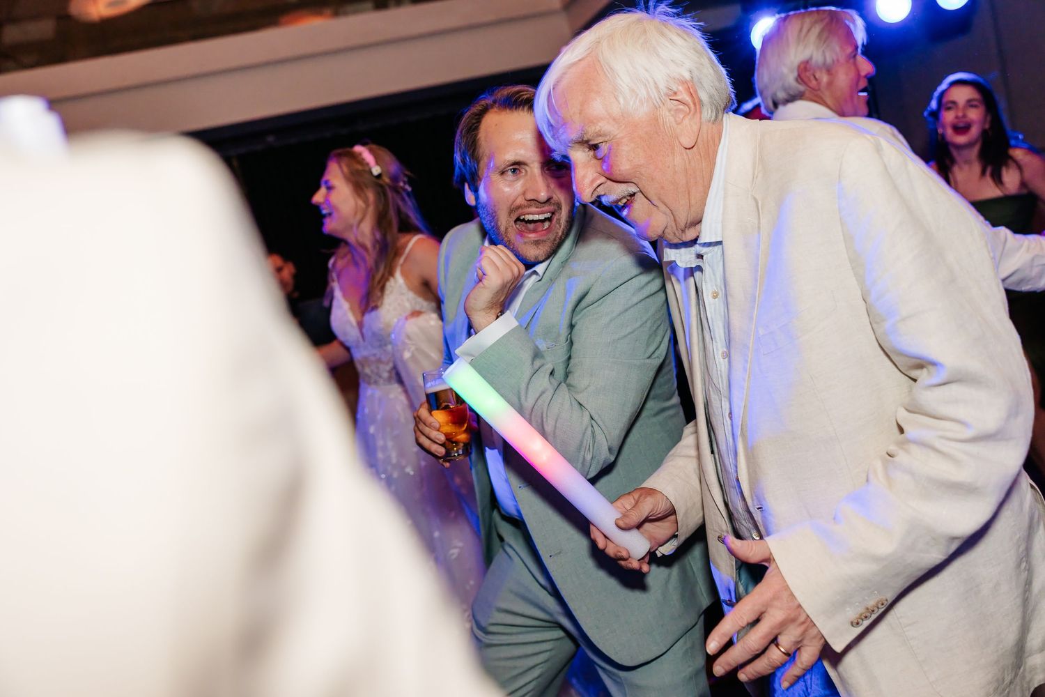 Elderly man dances energetically with guests under colorful lights at lively wedding reception party.