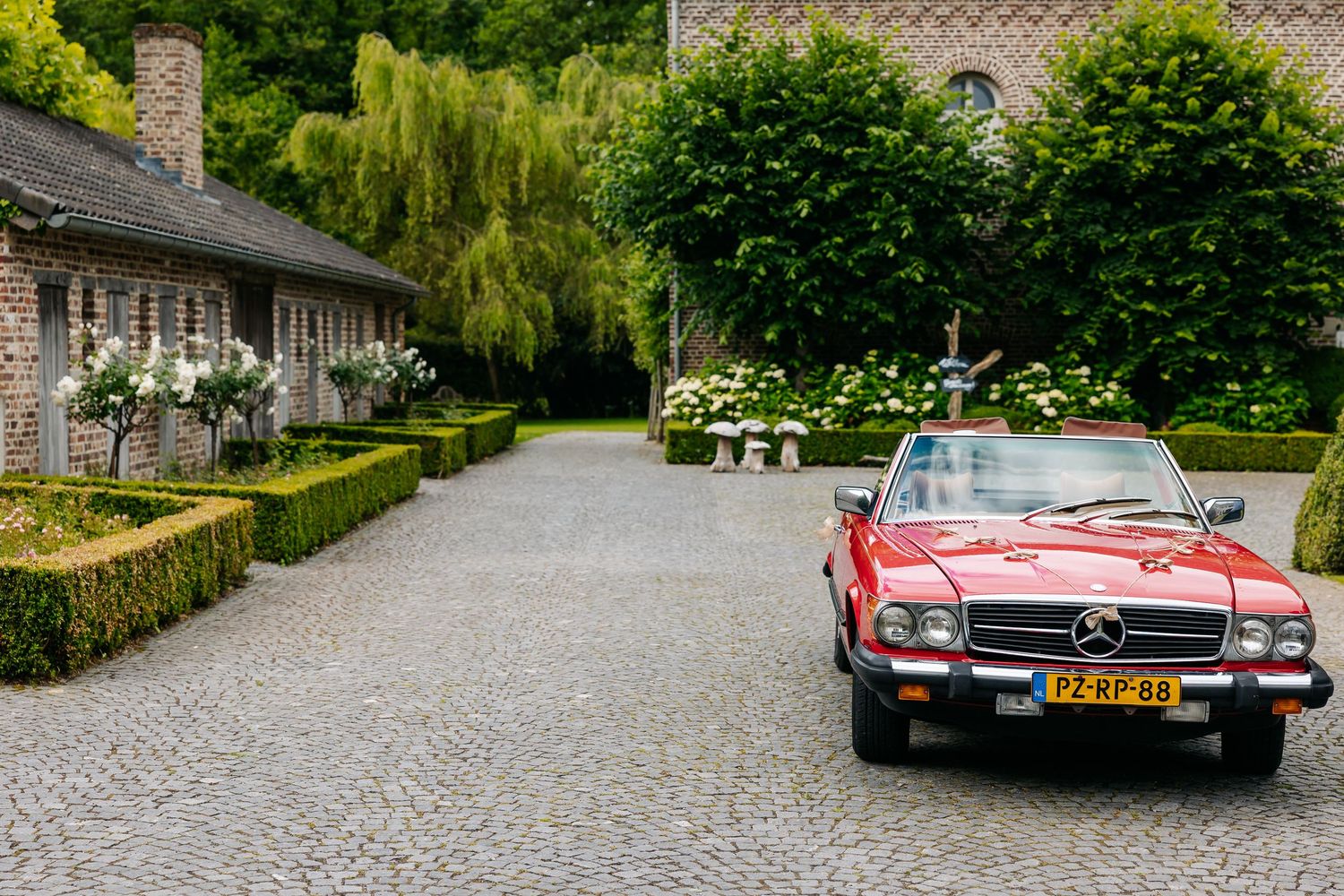 Red vintage Mercedes-Benz convertible parked on cobblestone driveway surrounded by lush green gardens.