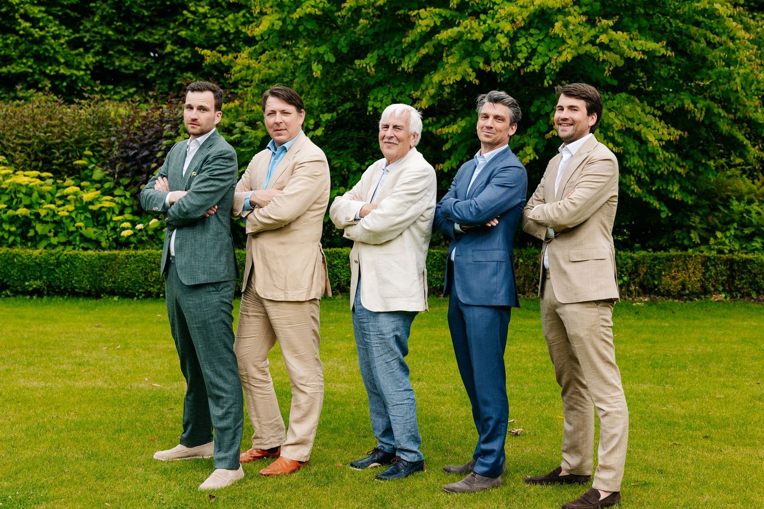 Five smartly dressed men in suits pose with arms crossed for a group photo on green lawn.