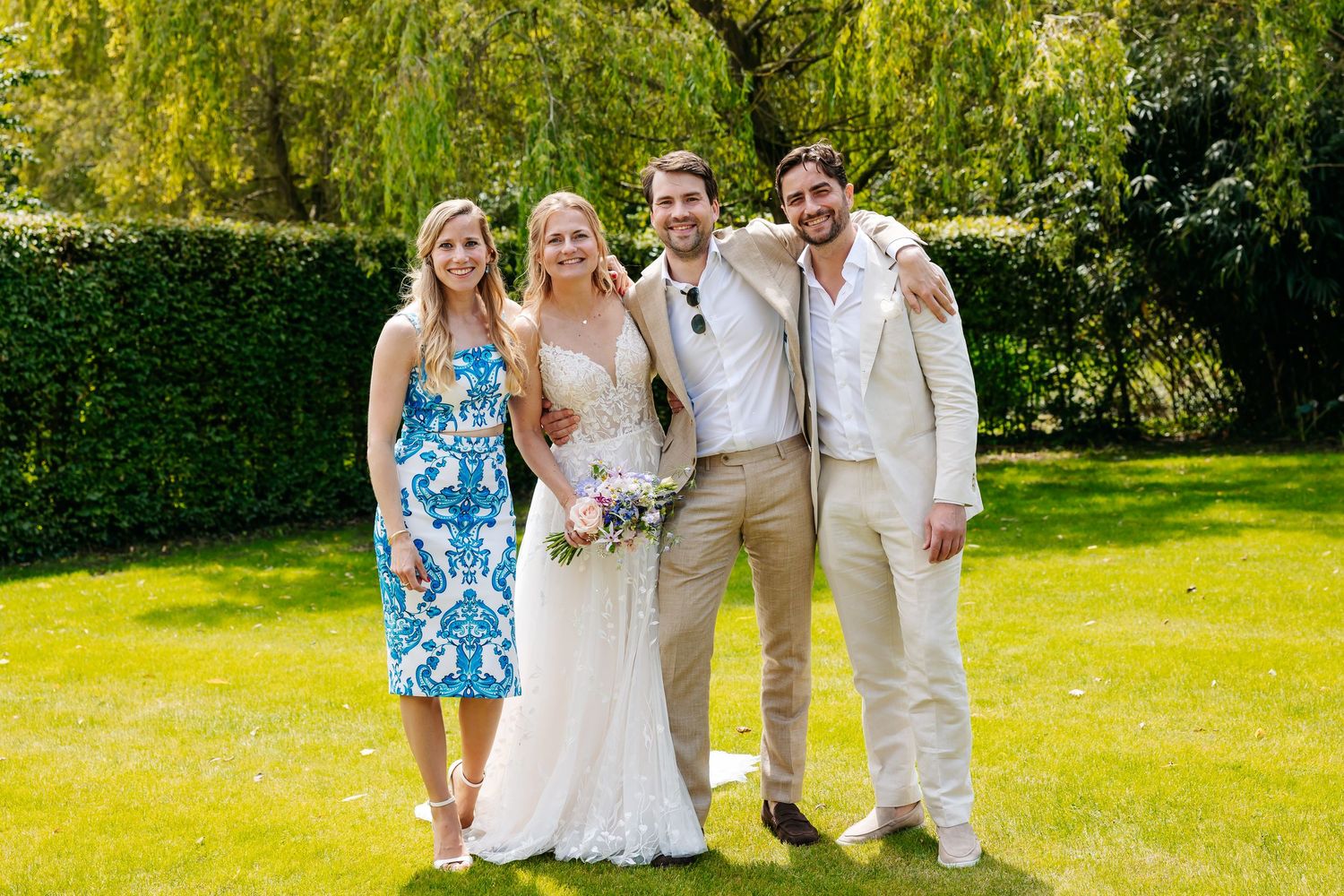 Bride and groom posing with two guests on green lawn, bride holding pink bouquet, groom in beige suit.