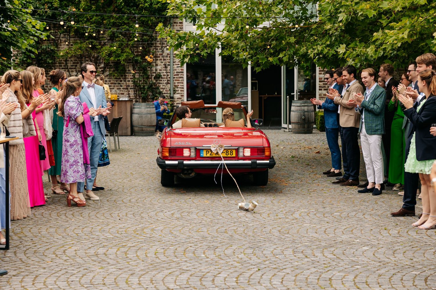 Newlyweds departing in a vintage red convertible as wedding guests cheer and applaud on both sides.