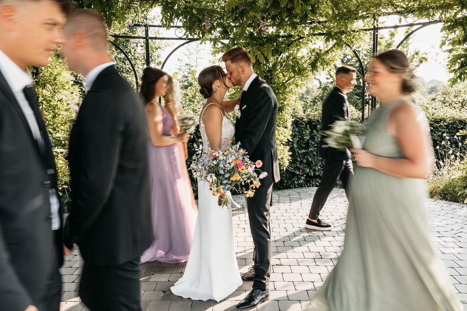 Bride and groom sharing a tender kiss surrounded by wedding party guests under a lush green arbor.