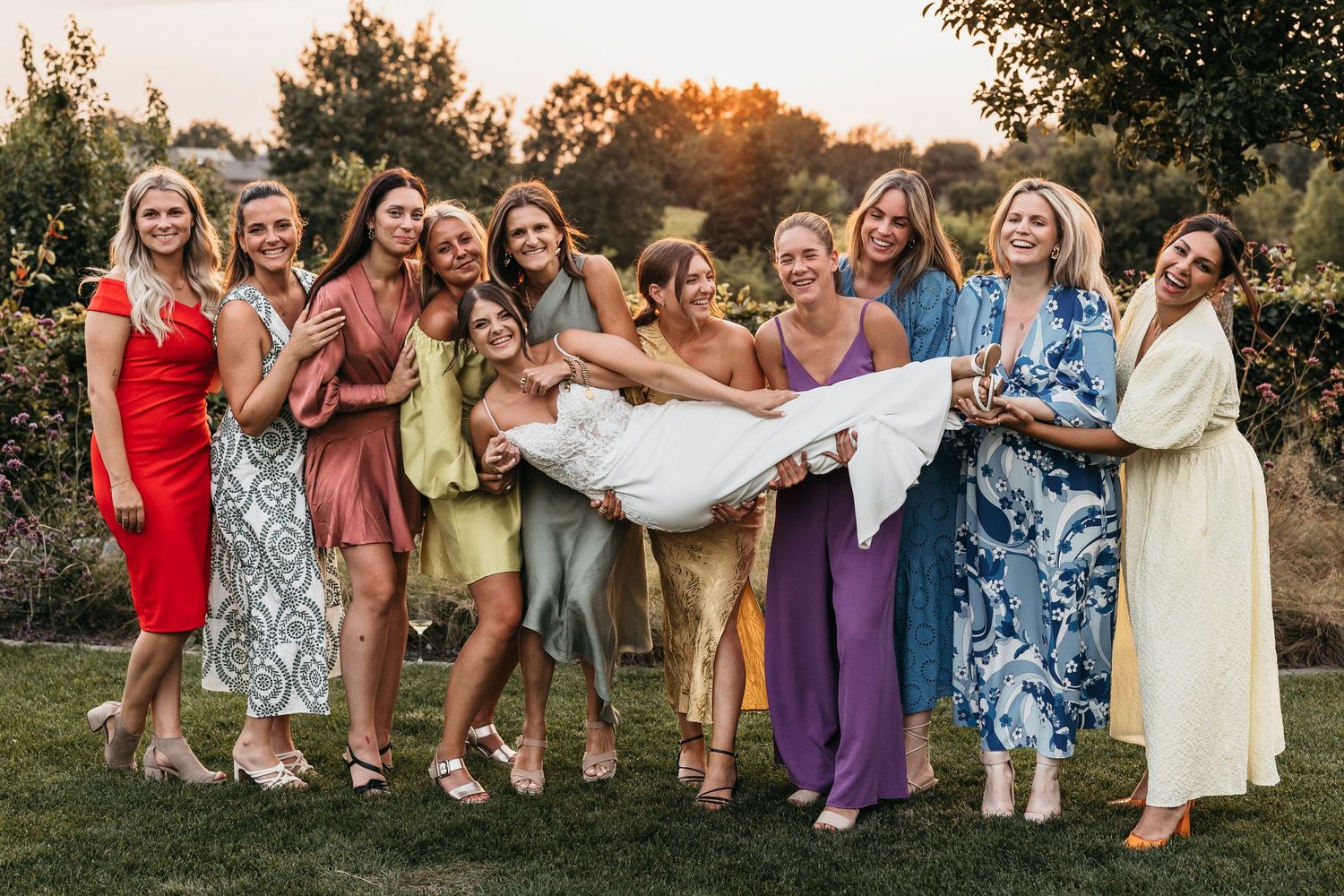 Bridesmaids in colorful dresses lifting smiling bride in white wedding gown outdoors.