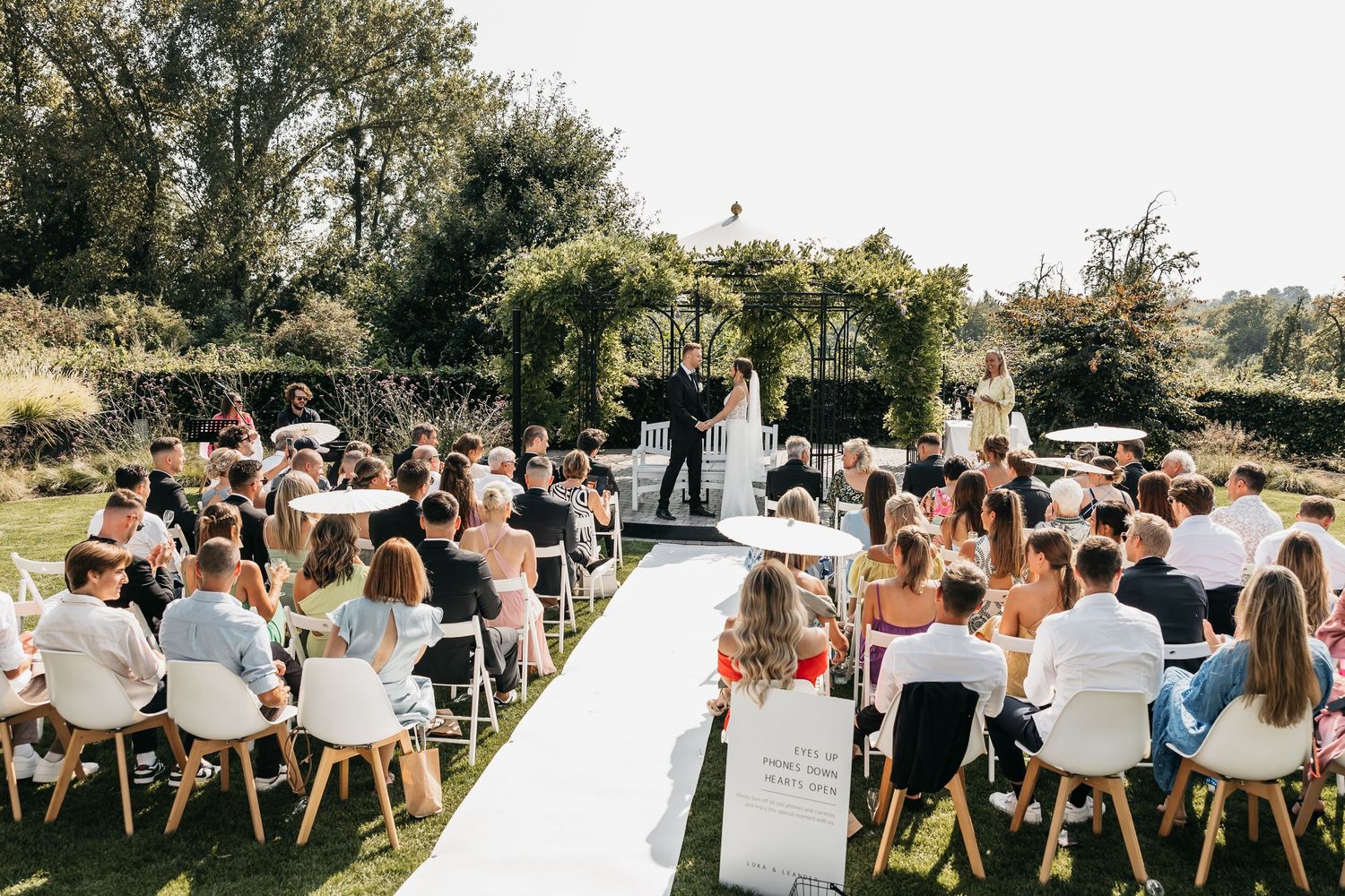 Wedding guests seated outdoors watching couple exchange vows under a floral arbor in a sunny garden.