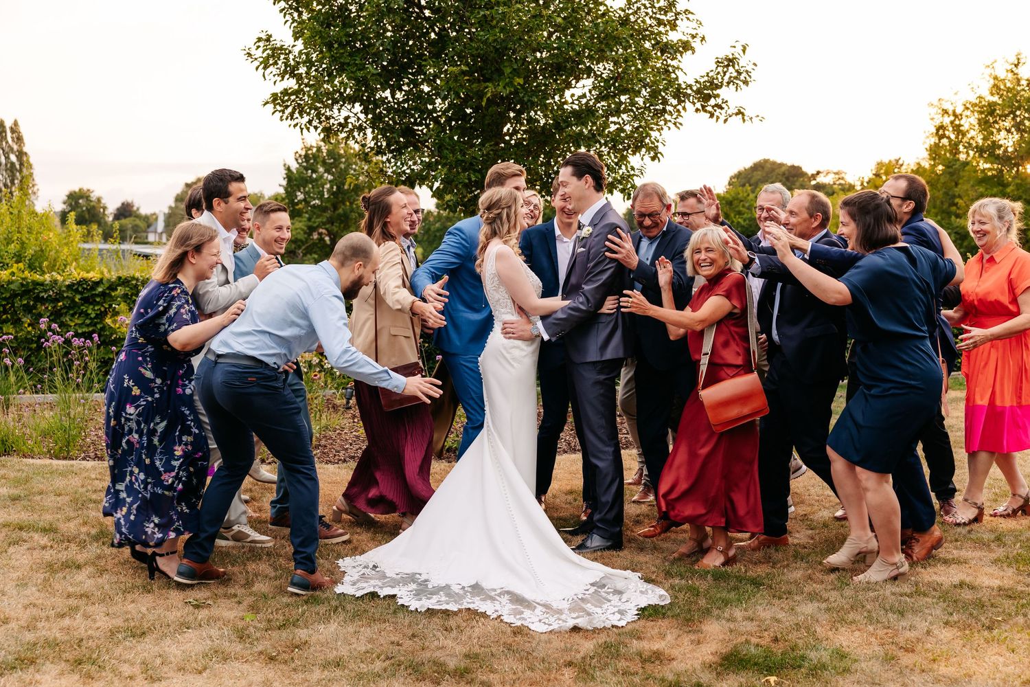 Joyful wedding party surrounds bride and groom in a fun outdoor group celebration photo.