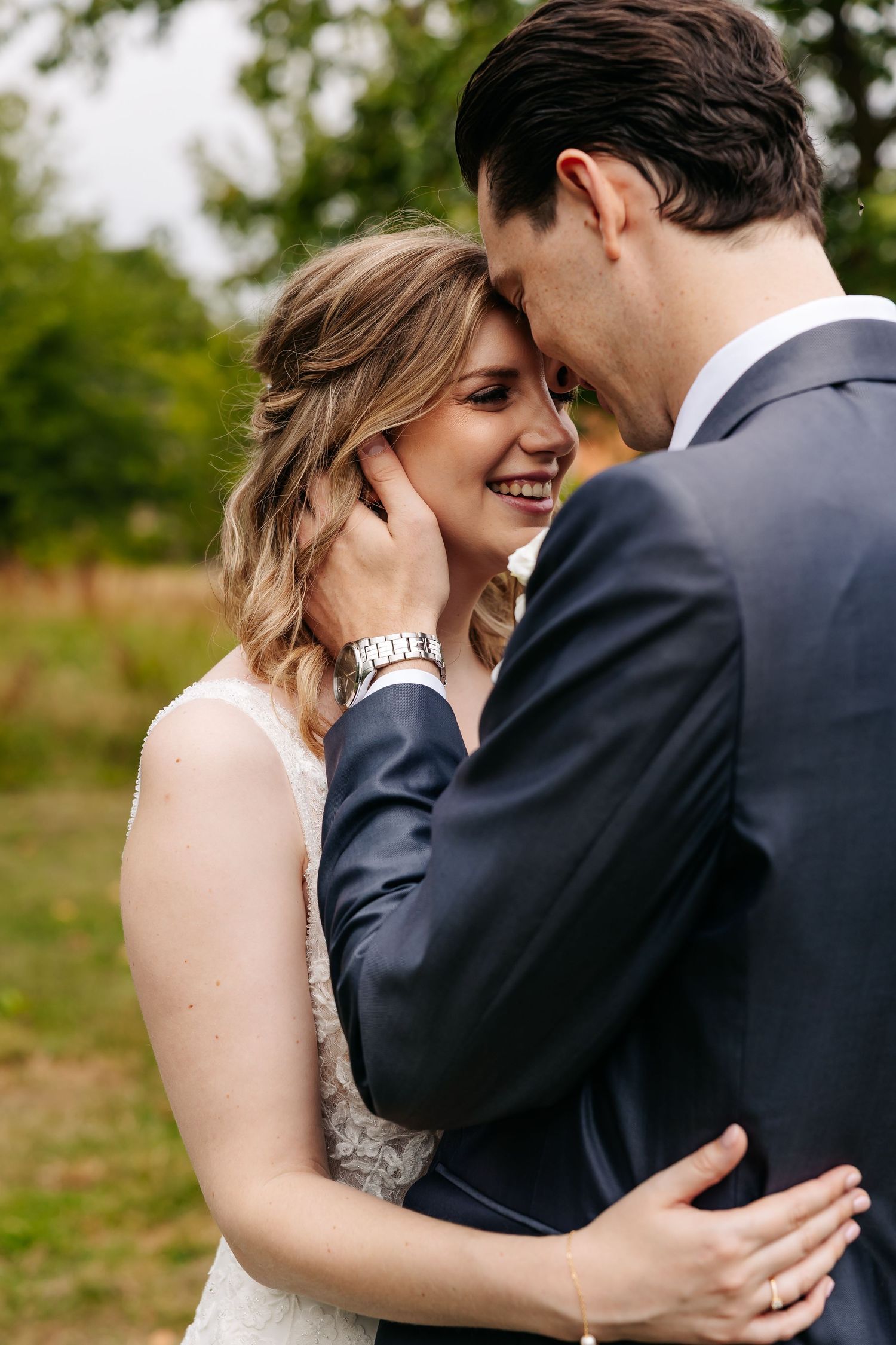 Bride laughing joyfully as groom gently cups her face outdoors with lush green trees in the background.