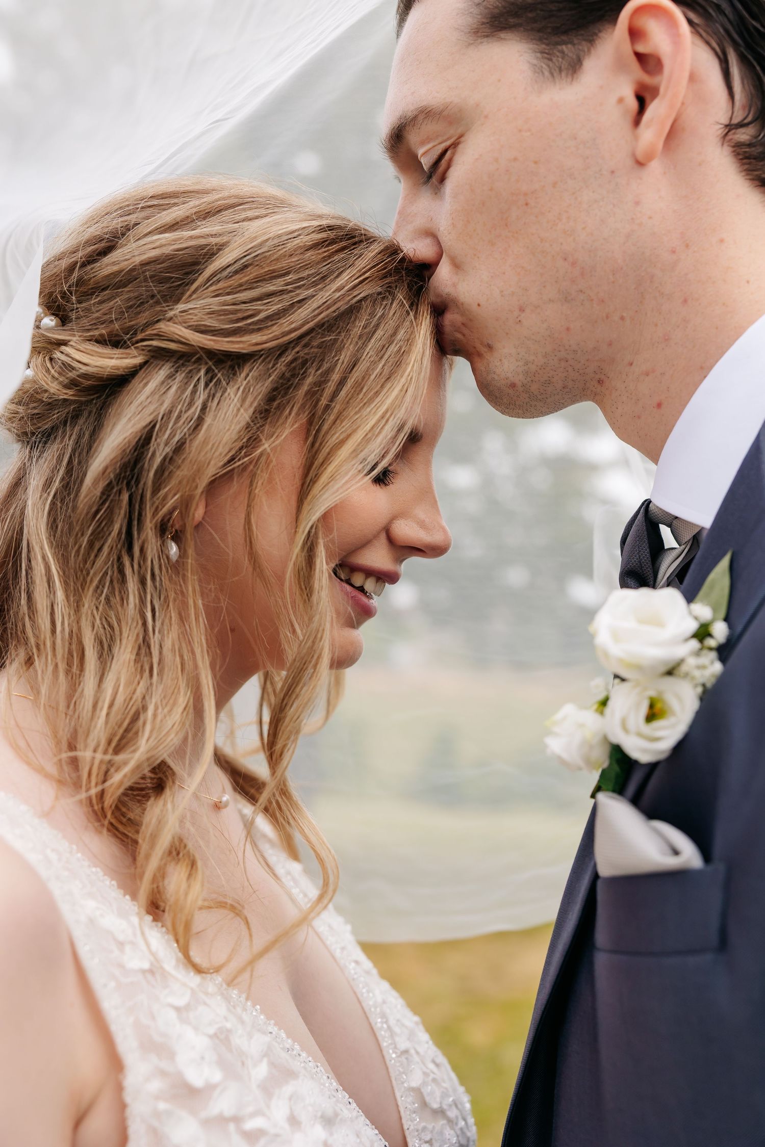 Groom kisses laughing brides forehead under a veil, showcasing her elegant updo and lace gown.