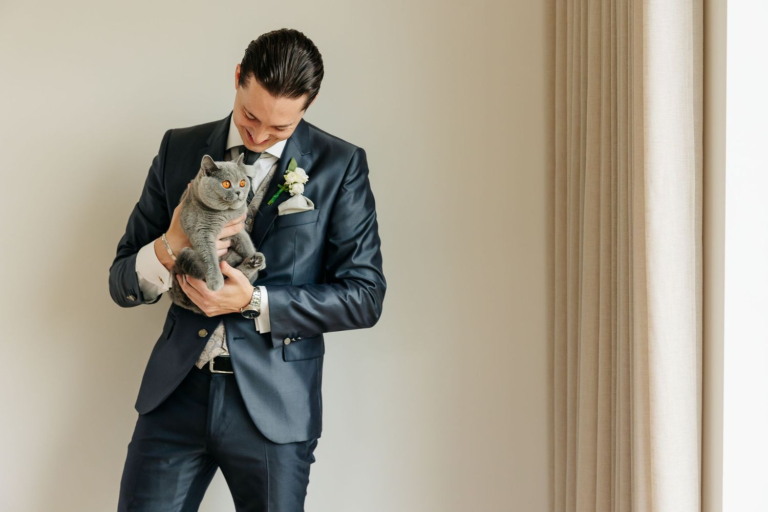 Dapper groom in navy suit with boutonniere holds a gray British Shorthair cat near window.