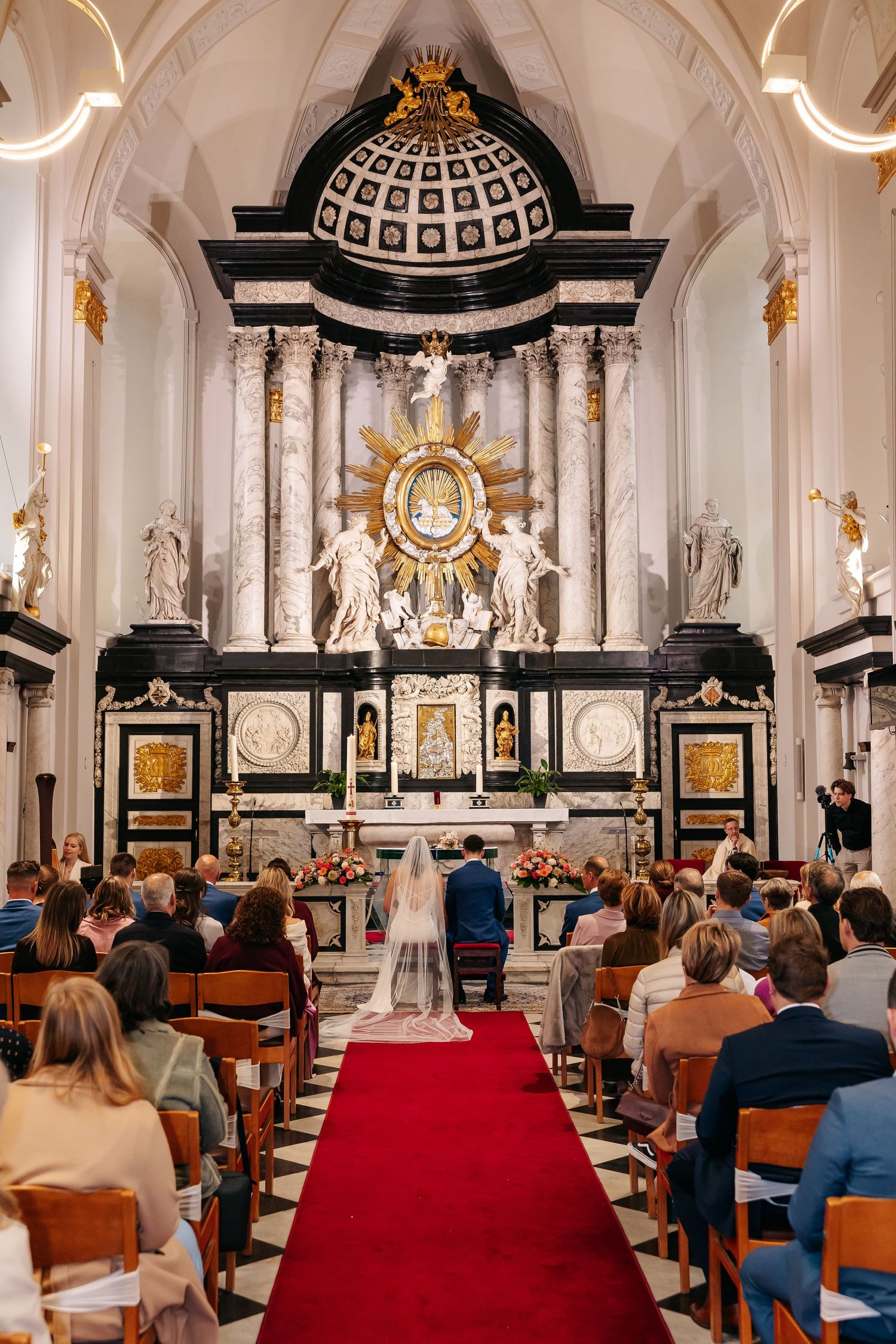 Wide view of elegant church wedding ceremony with guests seated along red carpet aisle.