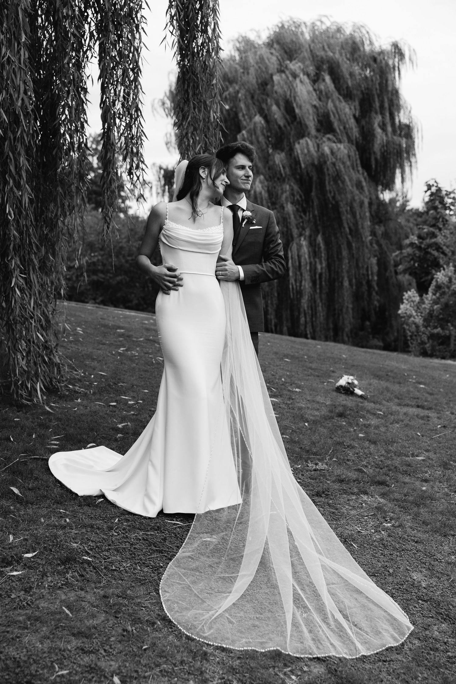 Black and white wedding portrait of couple embracing under weeping willow, bride's long veil trailing.