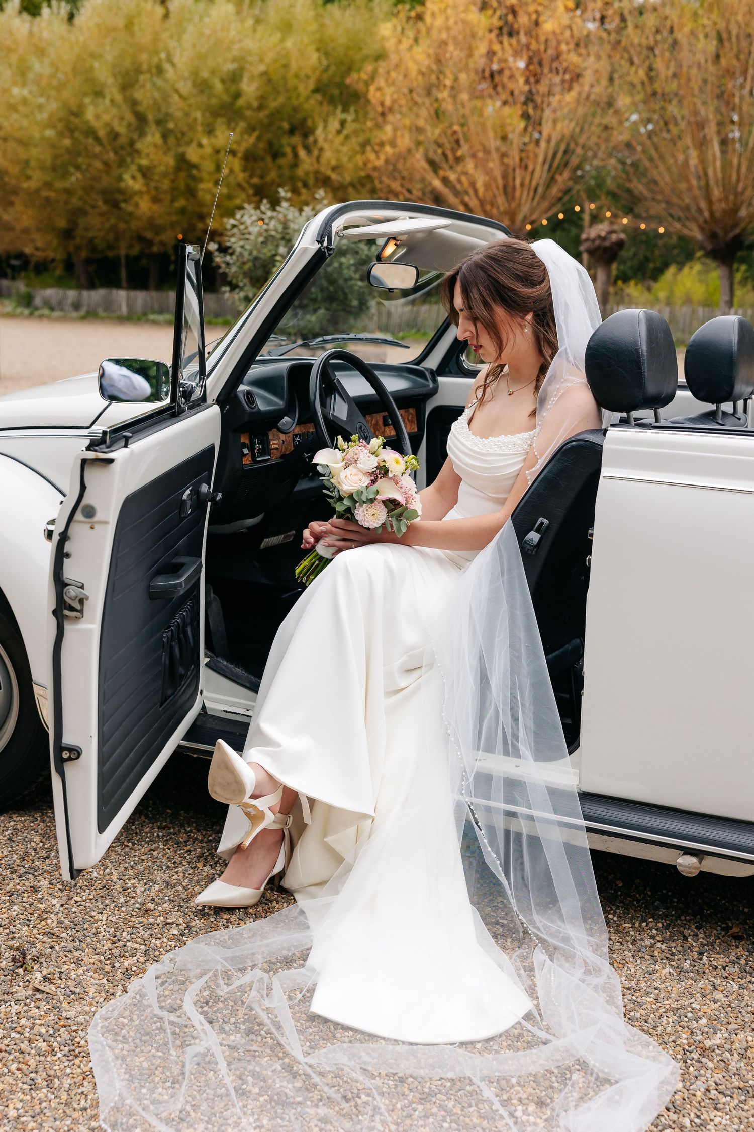 Bride in white wedding dress with long veil holding bouquet seated in a classic white vintage convertible car.