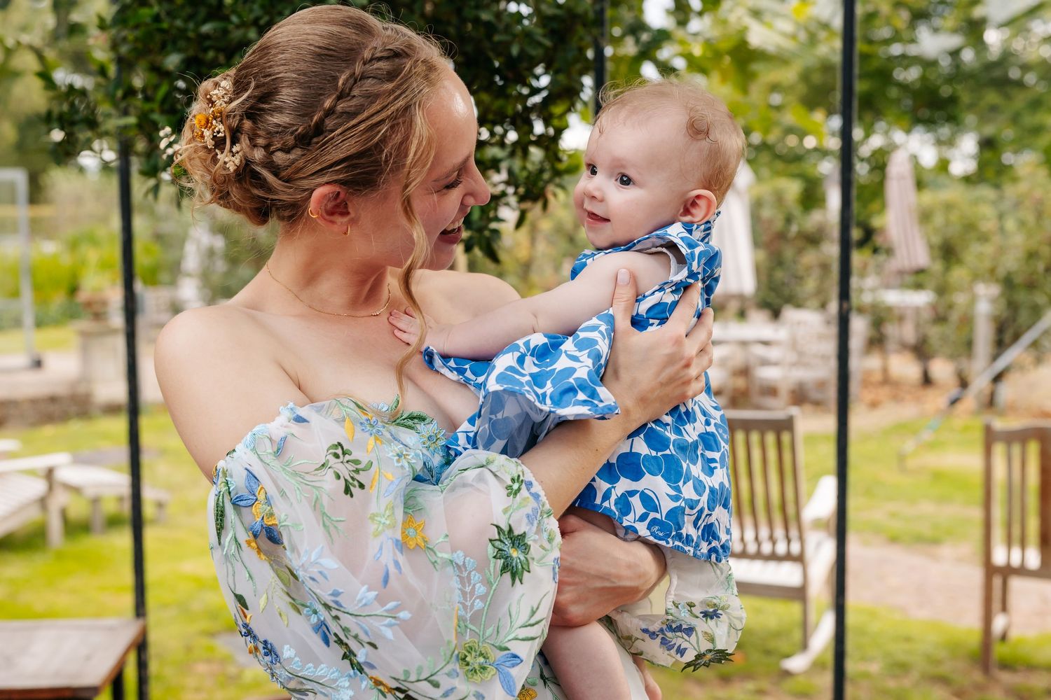 A smiling mother in a floral dress holds her baby girl in a blue outfit outdoors.