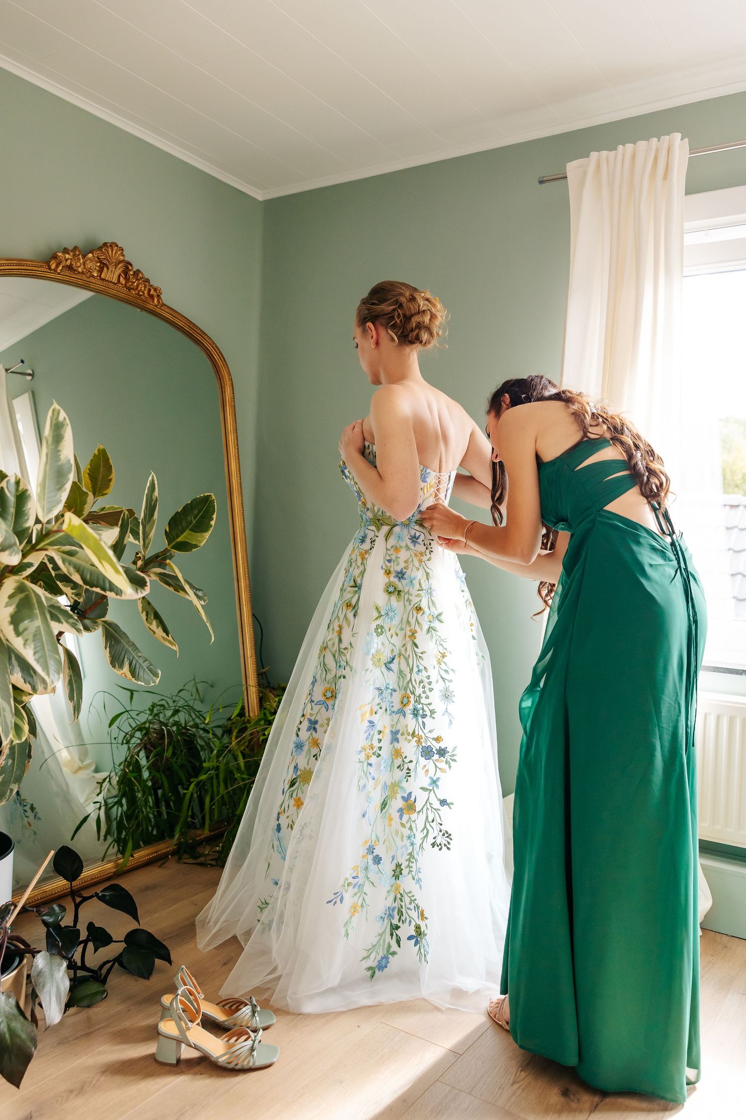 Bridesmaid in green dress fastening floral wedding gown on bride near gold ornate mirror.