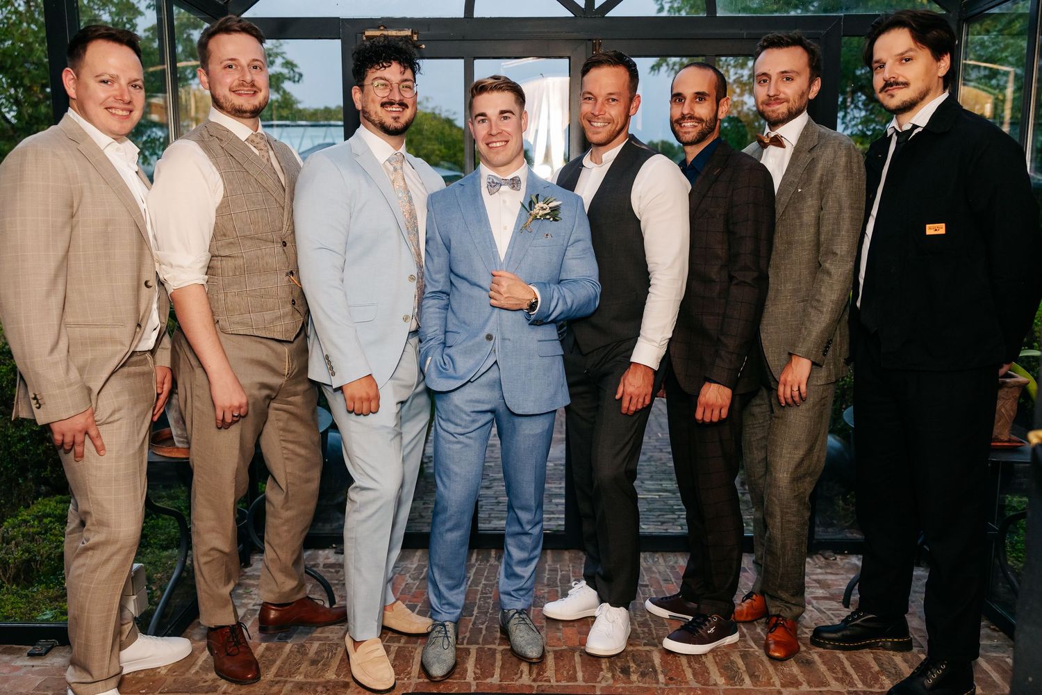 Groom poses with groomsmen in stylish suits inside a glass greenhouse wedding venue.