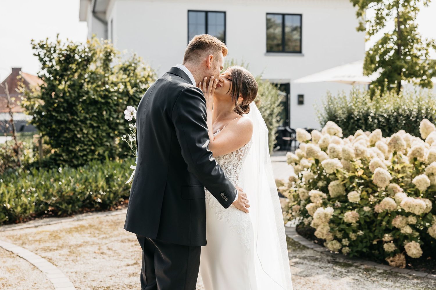 Wedding couple shares intimate kiss outdoors beside blooming white hydrangea bushes.