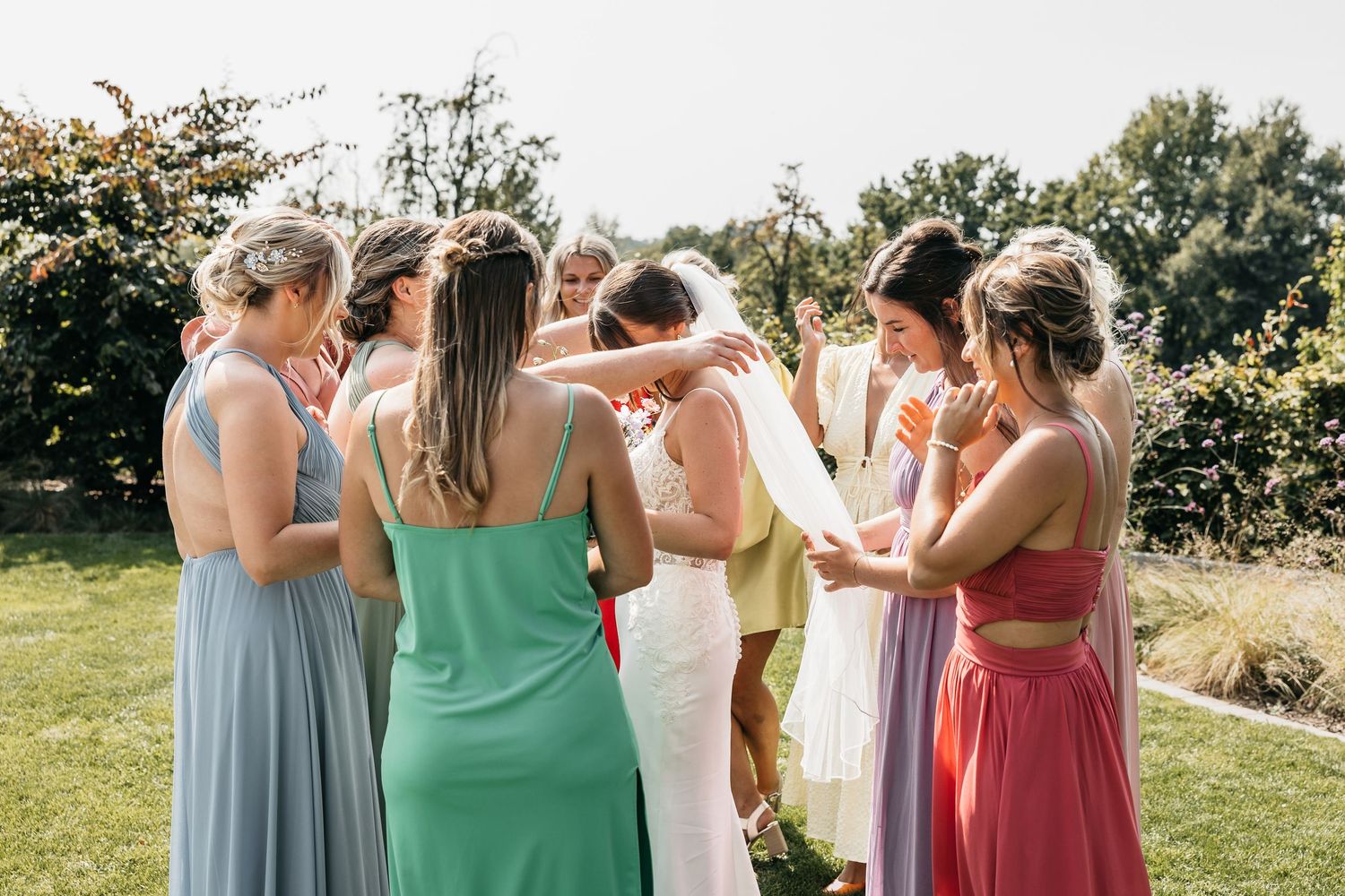 Bride in white gown shares heartfelt group hug with bridesmaids in colorful pastel dresses at outdoor wedding.