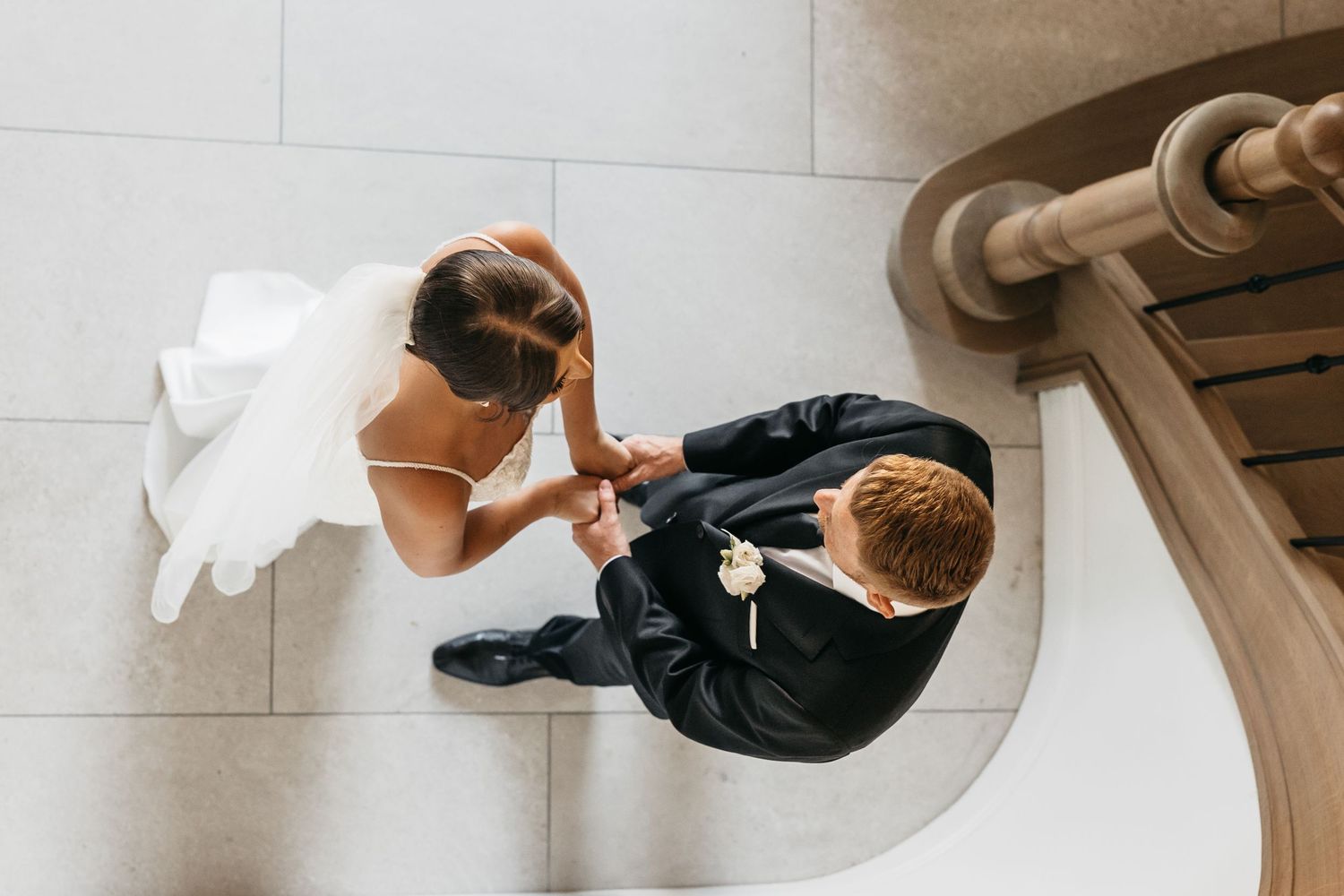 Aerial view of bride and groom holding hands at base of staircase on wedding day.