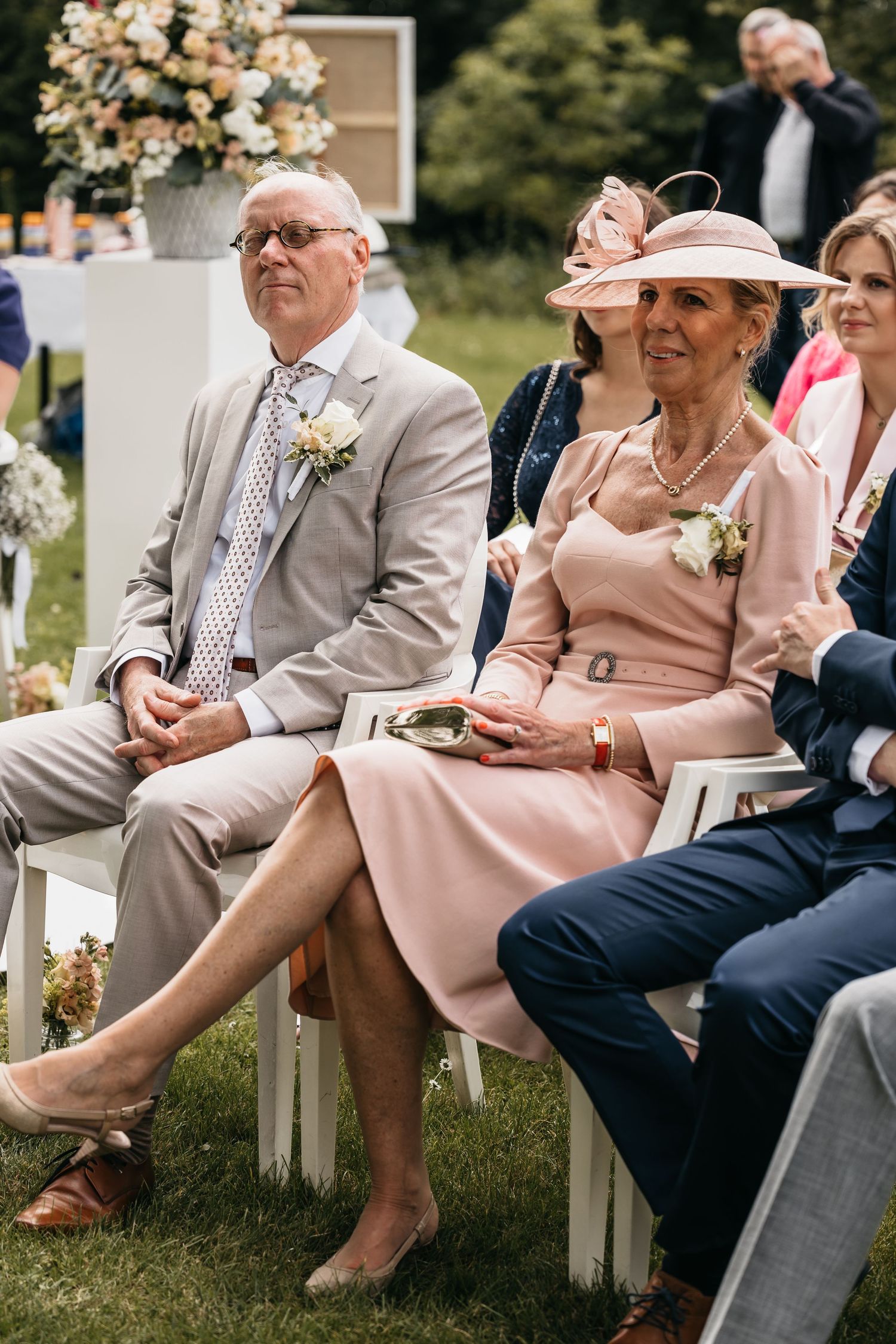 Wedding guests in elegant attire, including a woman in a pink dress and hat, watch the ceremony.