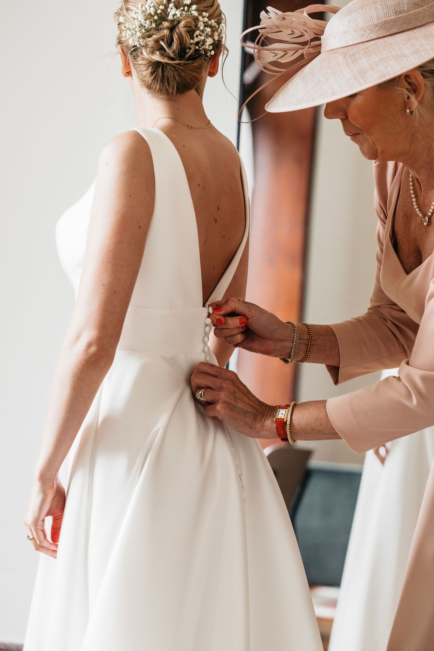 Mother in pink hat carefully buttoning the back of bride's sleek white wedding gown.