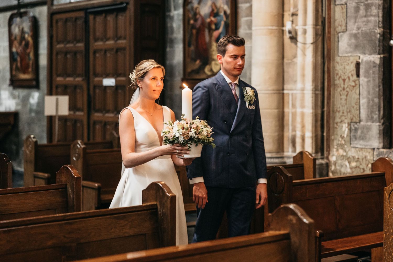 Bride holding bouquet walking with groom through historic stone church before wedding ceremony.