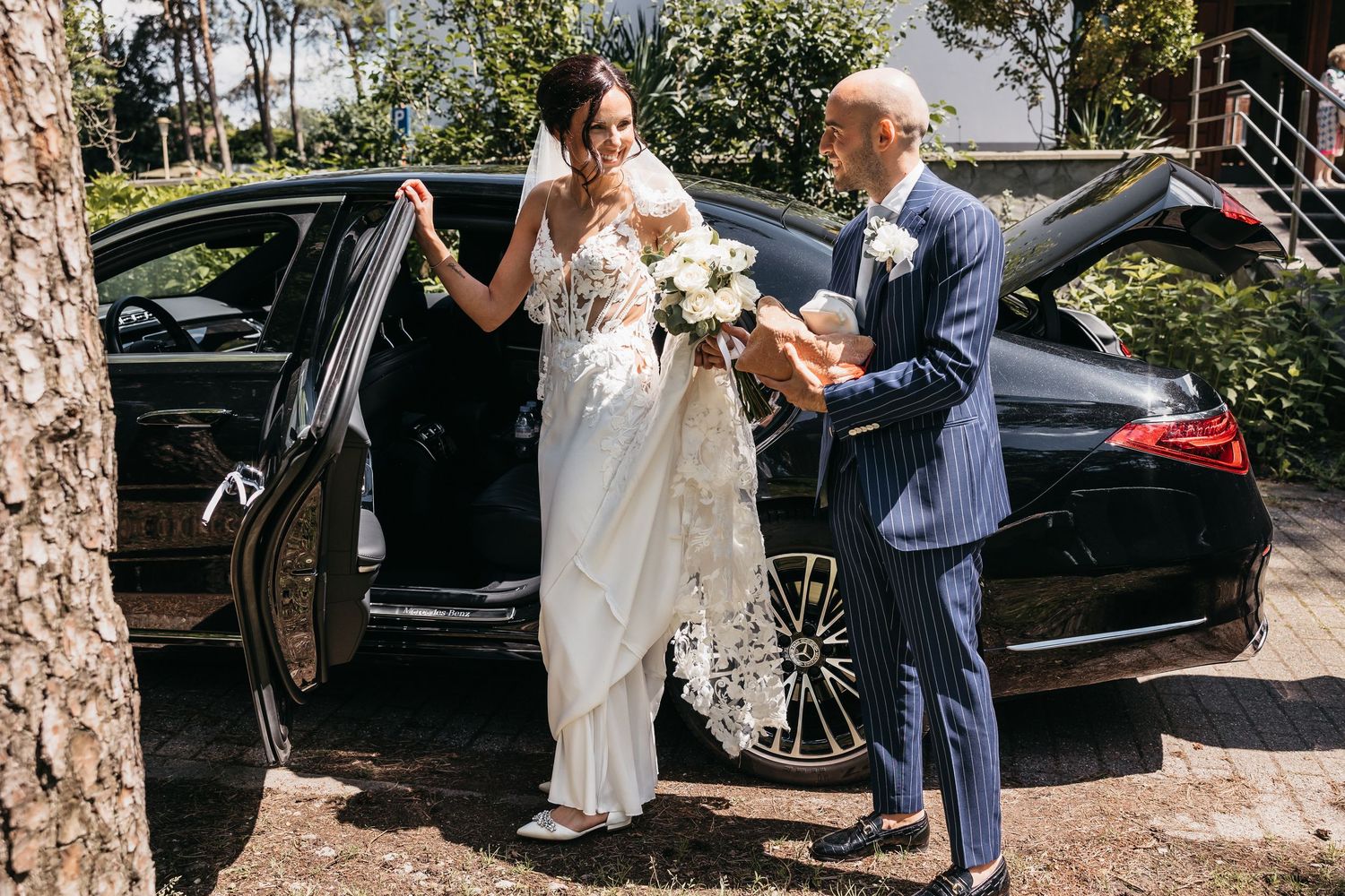 Bride in white gown arriving at wedding venue assisted by man in blue suit beside black car.