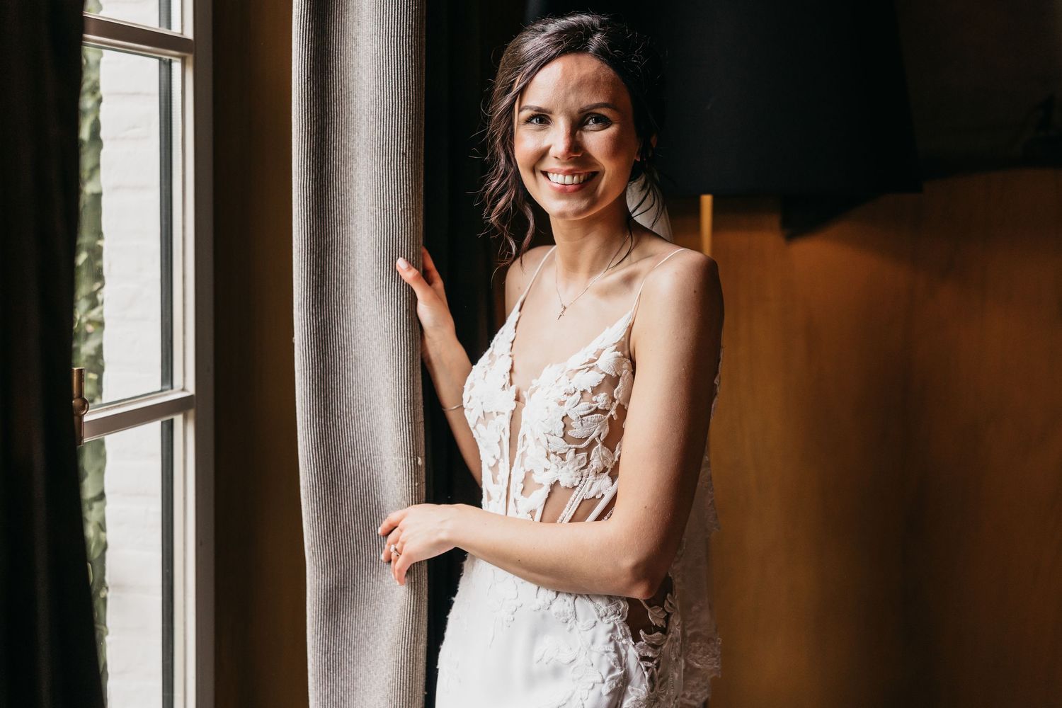 Joyful bride in a floral lace wedding gown smiles while standing near a sunlit window.