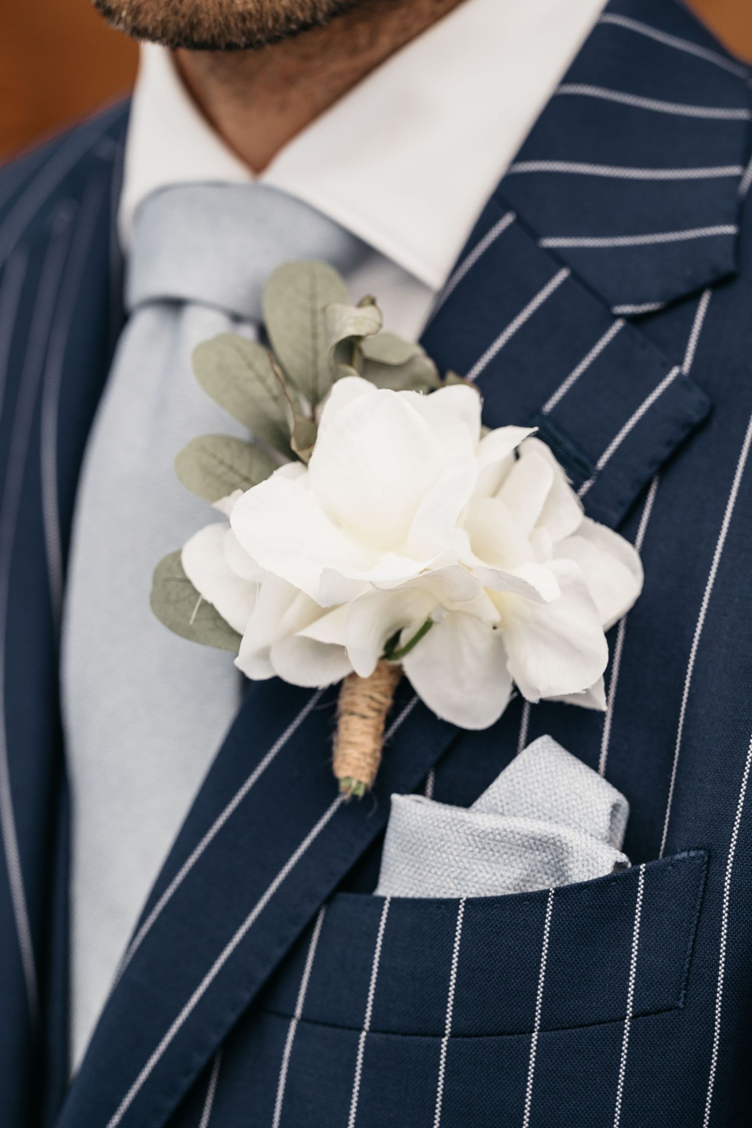 Close-up of white rose boutonniere with eucalyptus on navy pinstripe wedding suit jacket.