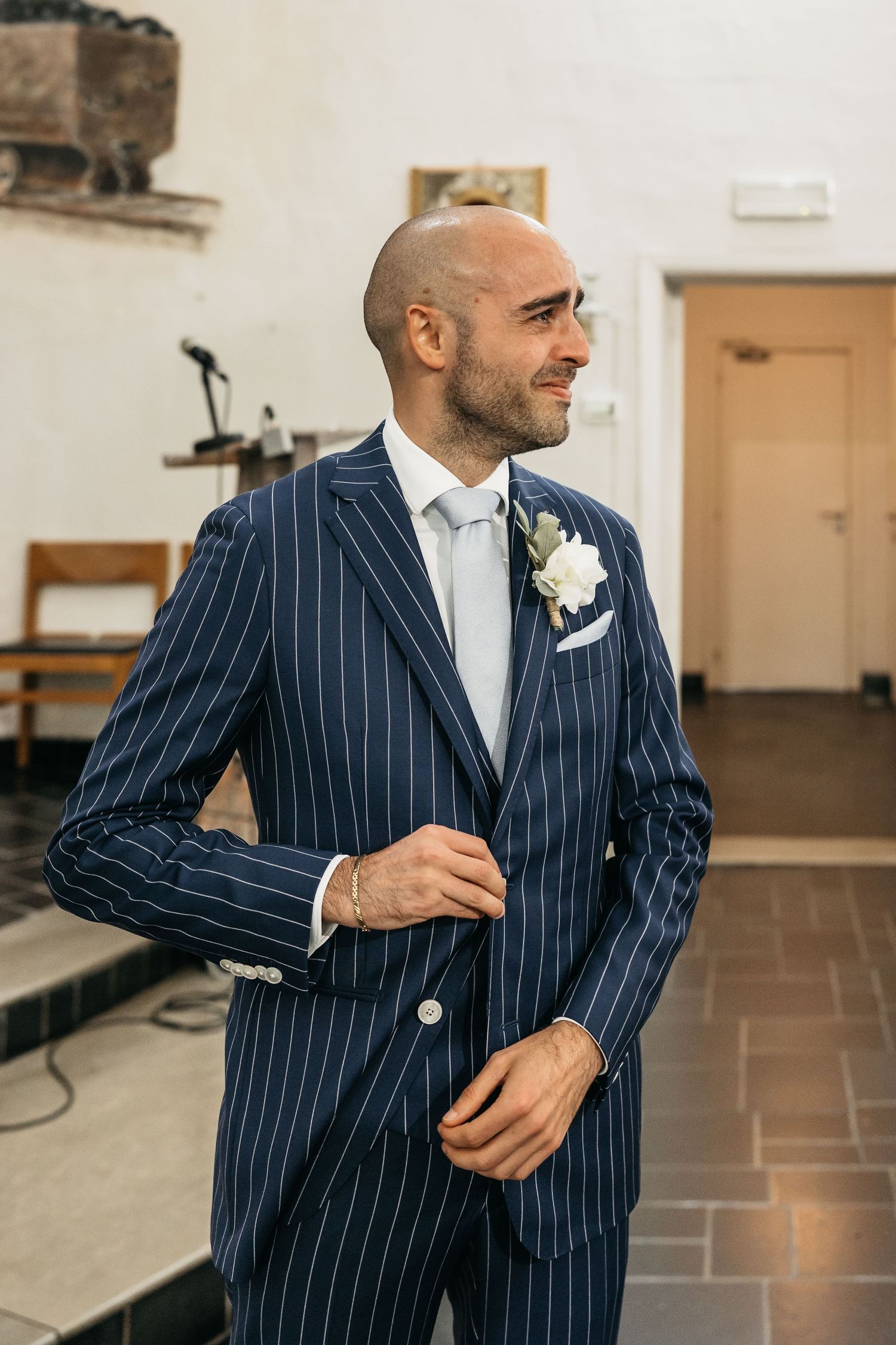 Dapper bald groom in navy pinstripe three-piece suit with white rose boutonniere adjusting his jacket in church.