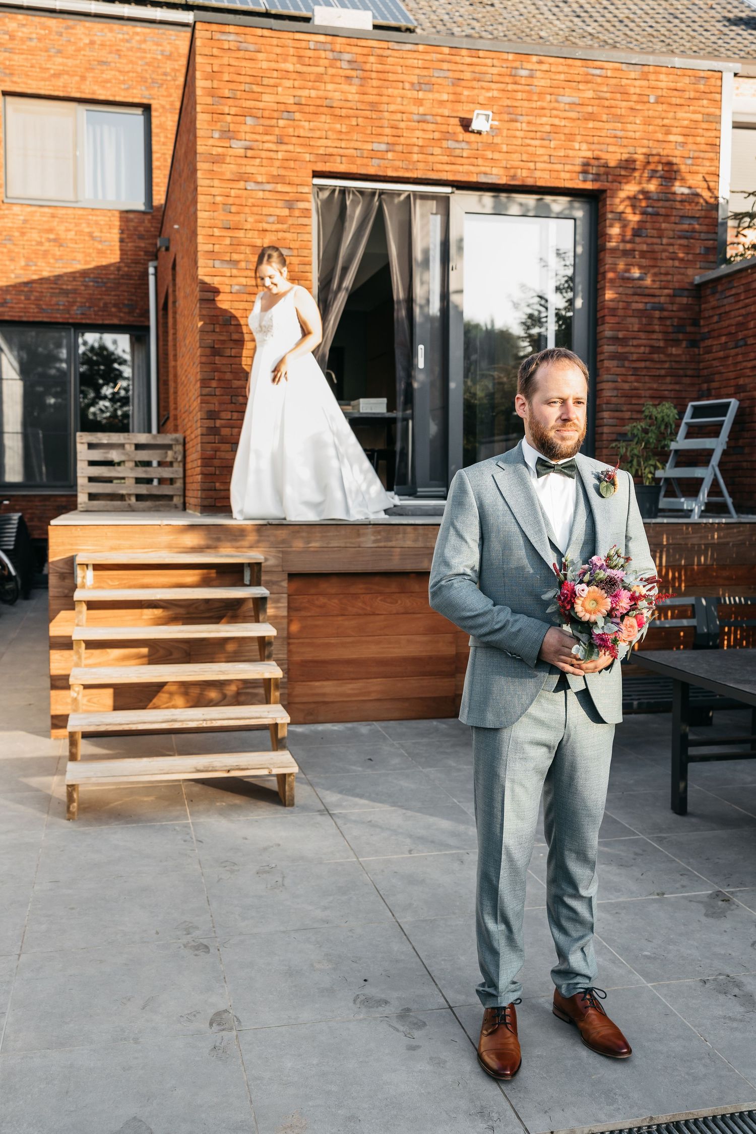 Bride in white gown on deck steps while groom holds bouquet in foreground at home.